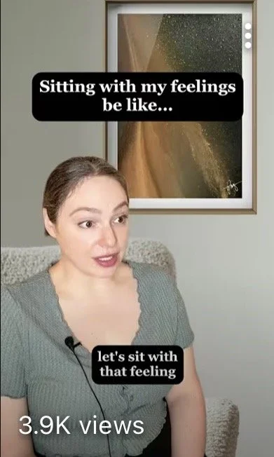 A woman with brown hair pulled back, sitting on a couch in front of an abstract painting. Text overlay says 'Sitting with my feelings be like... let's sit with that feeling.'