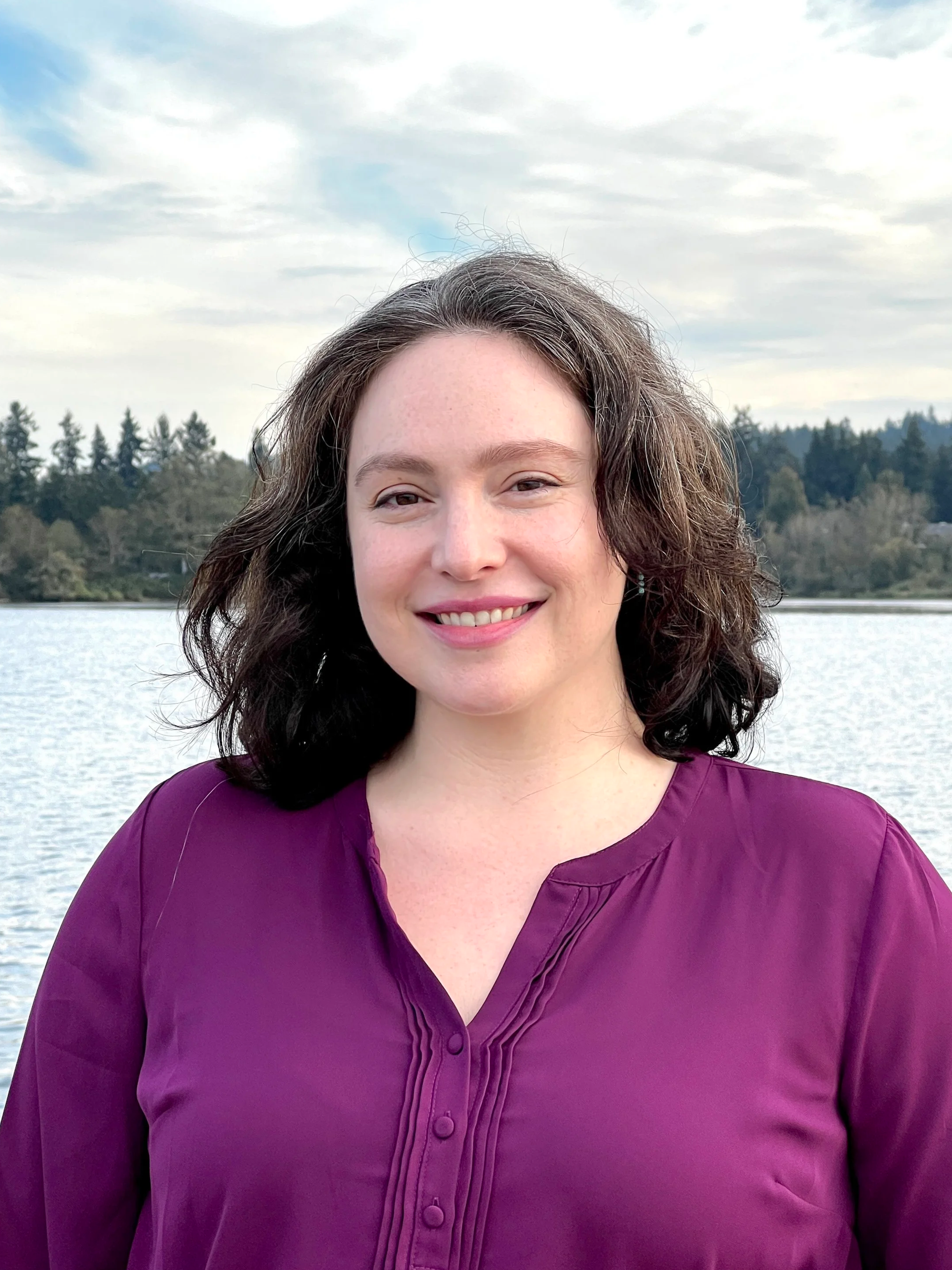 A woman with shoulder-length dark hair smiling outdoors near a body of water, with trees and a cloudy sky in the background.