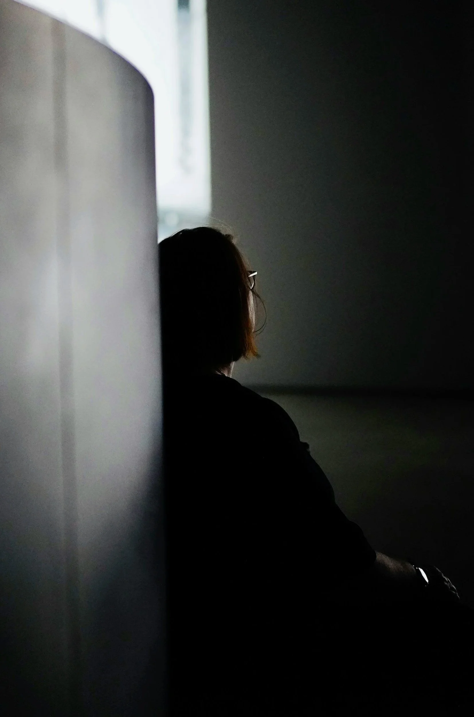 A person with shoulder-length hair and glasses sitting with a window providing soft natural light behind them.