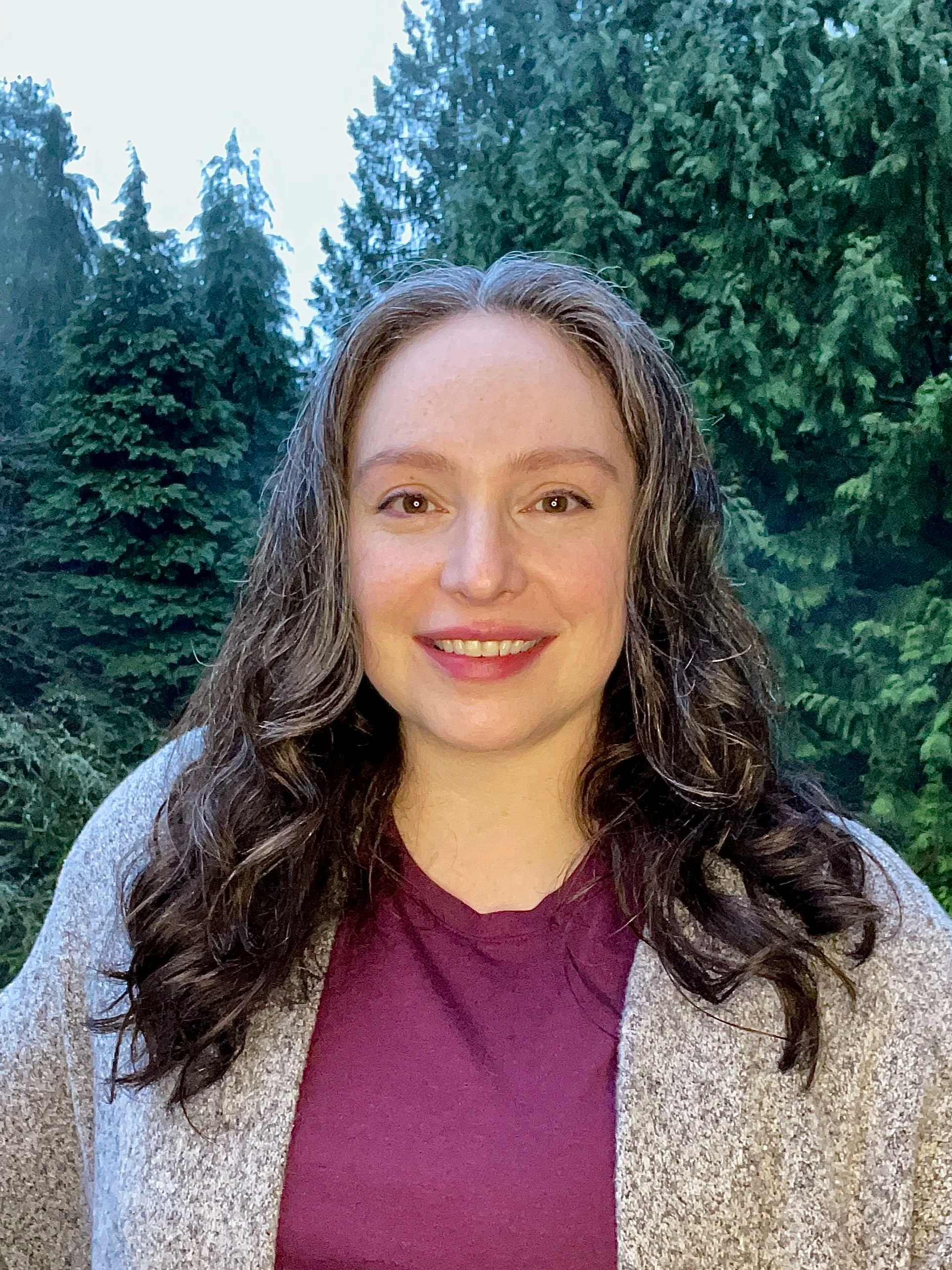 A woman with long, curly brown hair, smiling, outdoors with green trees in the background, wearing a purple top and a gray cardigan.
