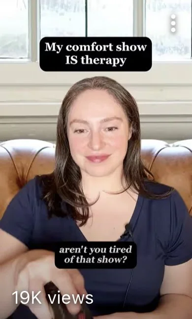 A woman with brown hair sitting on a leather couch in front of a window, with text overlay that says "My comfort show IS therapy" and "aren't you tired of that show?"