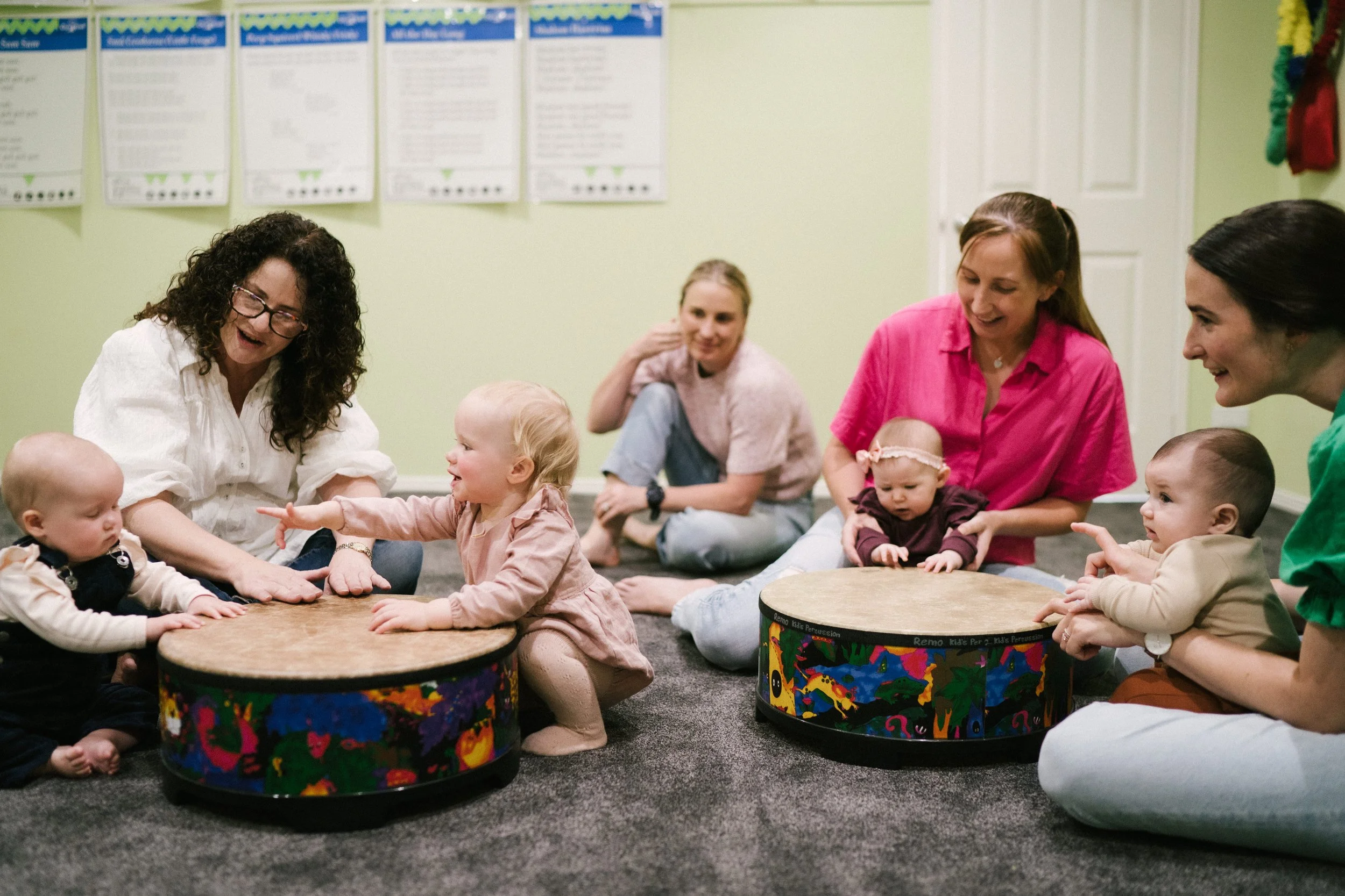 A group of women and babies sitting on a carpeted floor in a room, engaging in a playful activity with round drums. Two women are helping babies explore the drums while smiling and interacting with them.