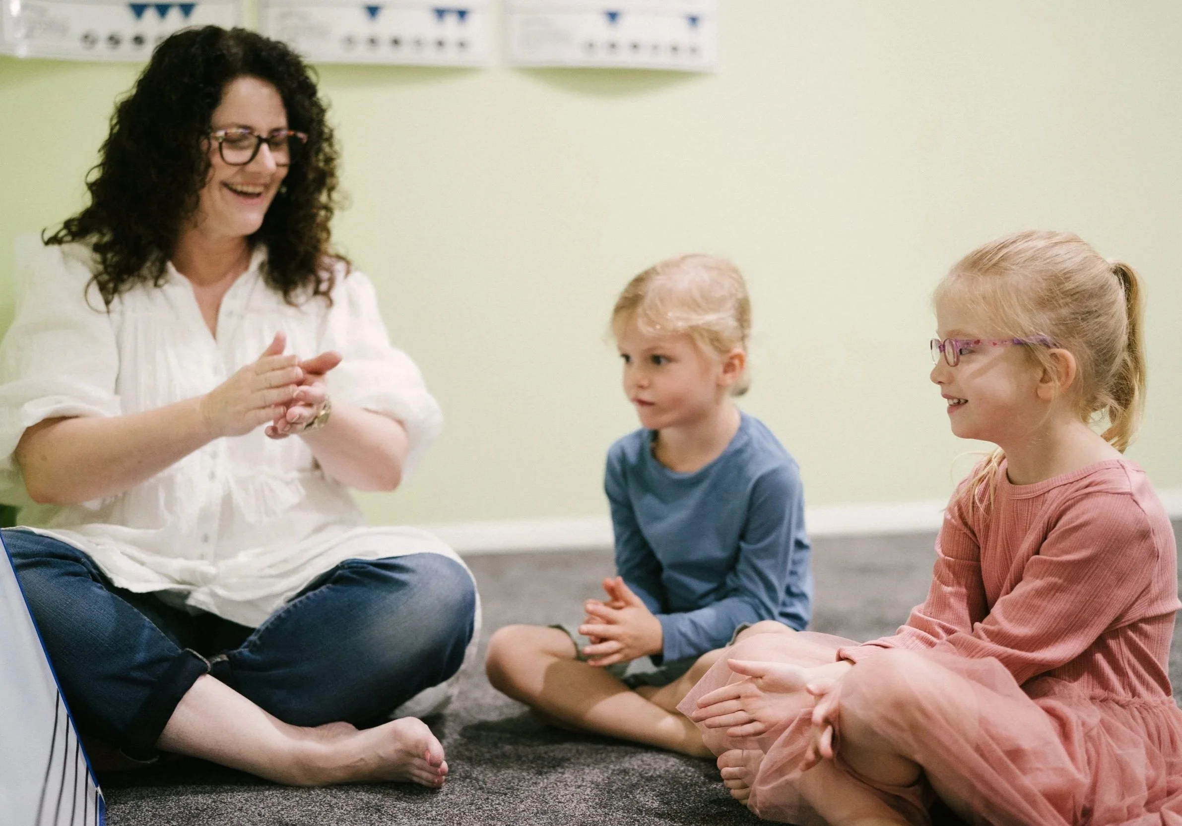 A woman with curly hair and glasses sitting cross-legged on the floor, smiling and clapping her hands, talking to two children, a boy in a blue shirt and a girl in a pink dress, who are sitting nearby and listening attentively.