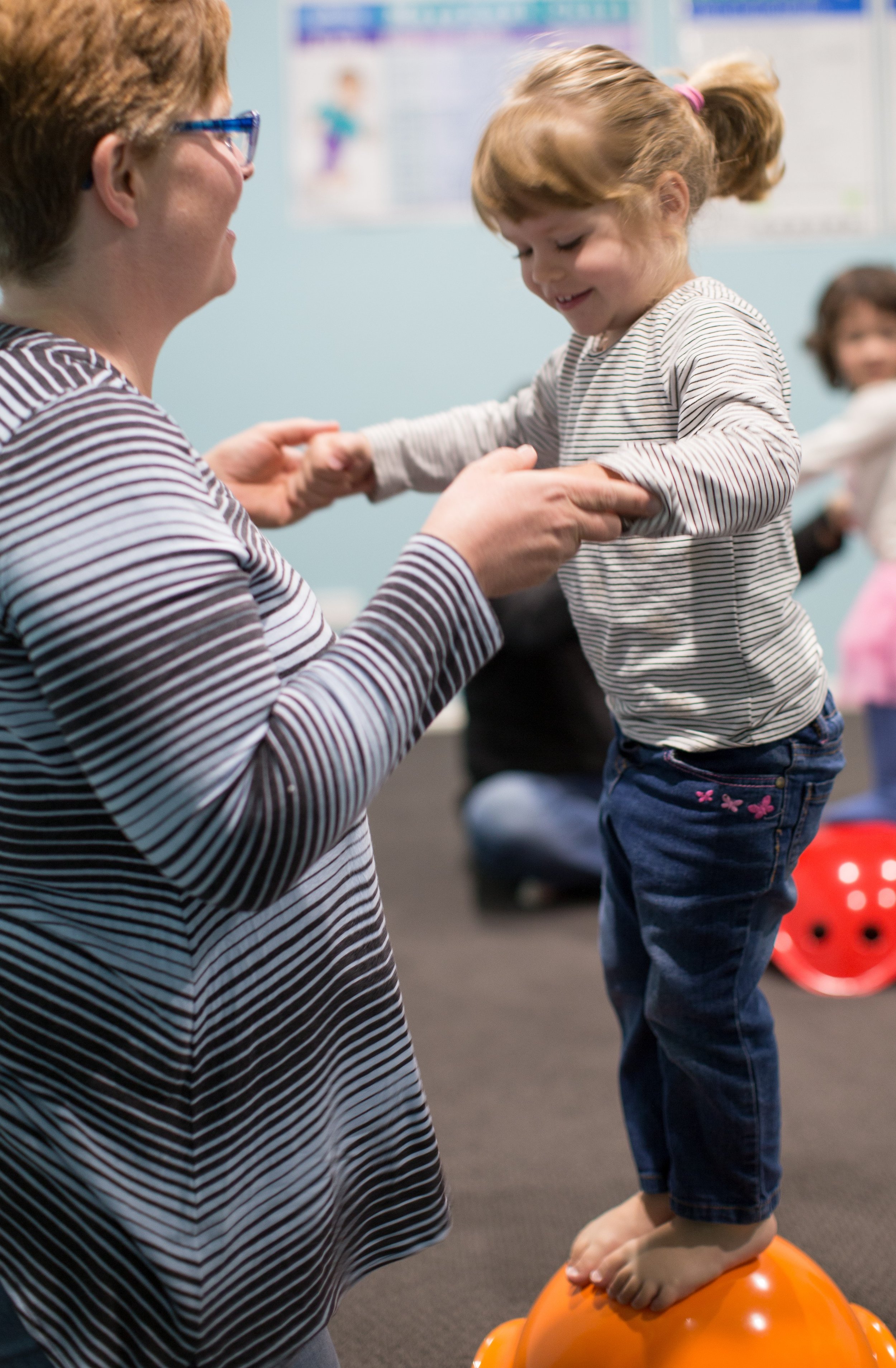 A young girl stands on an orange balance ball while holding hands with her mum, having fun during a music and movement class.