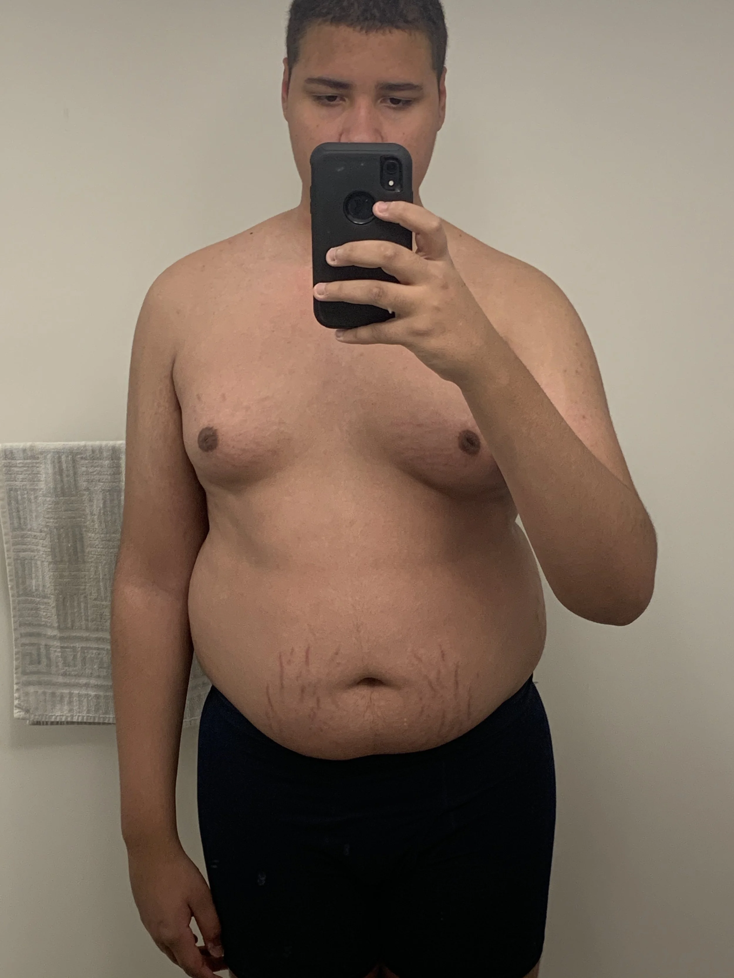 A shirtless man taking a mirror selfie in a room with a white wall and a towel rack visible in the background.