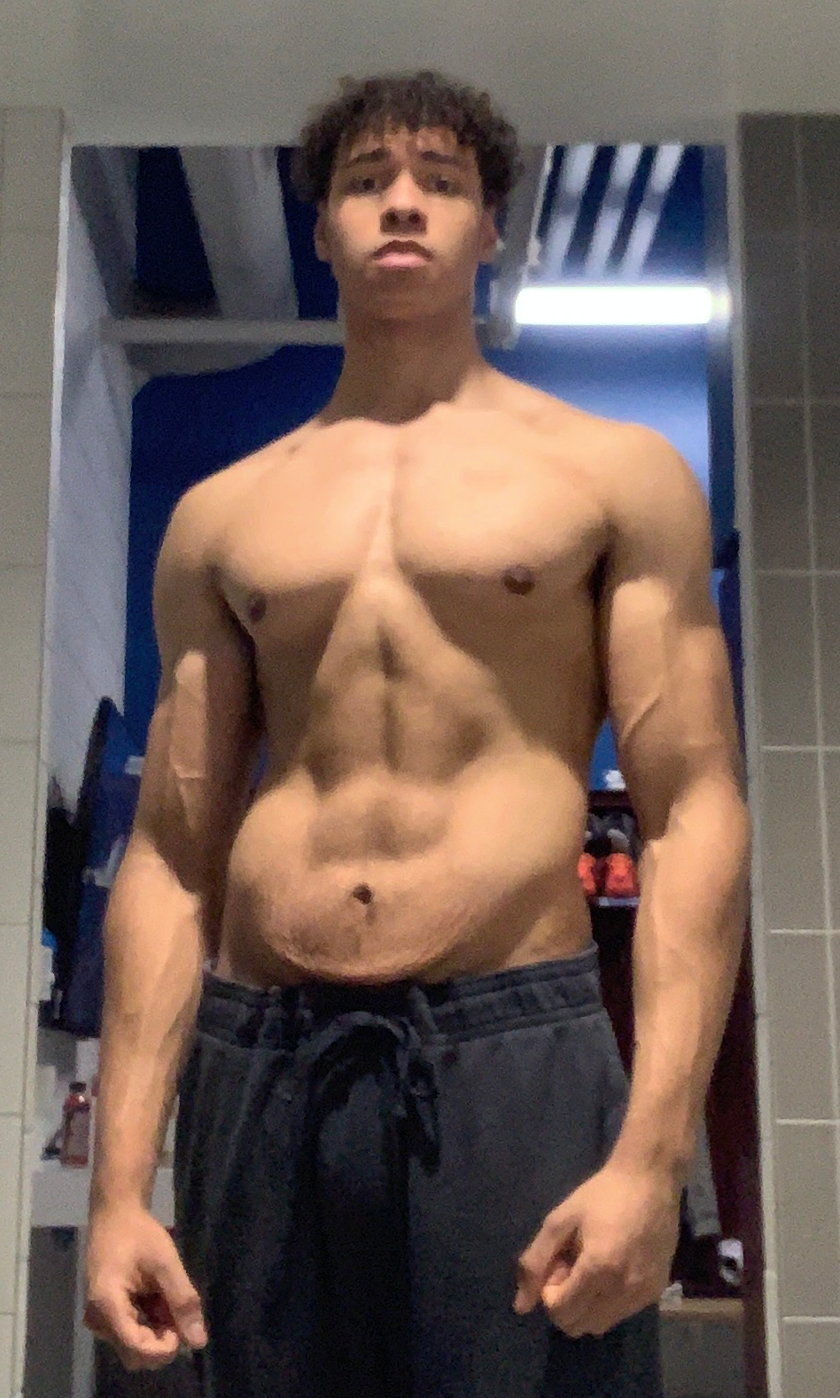 A shirtless young man with curly hair taking a mirror selfie in a bathroom.