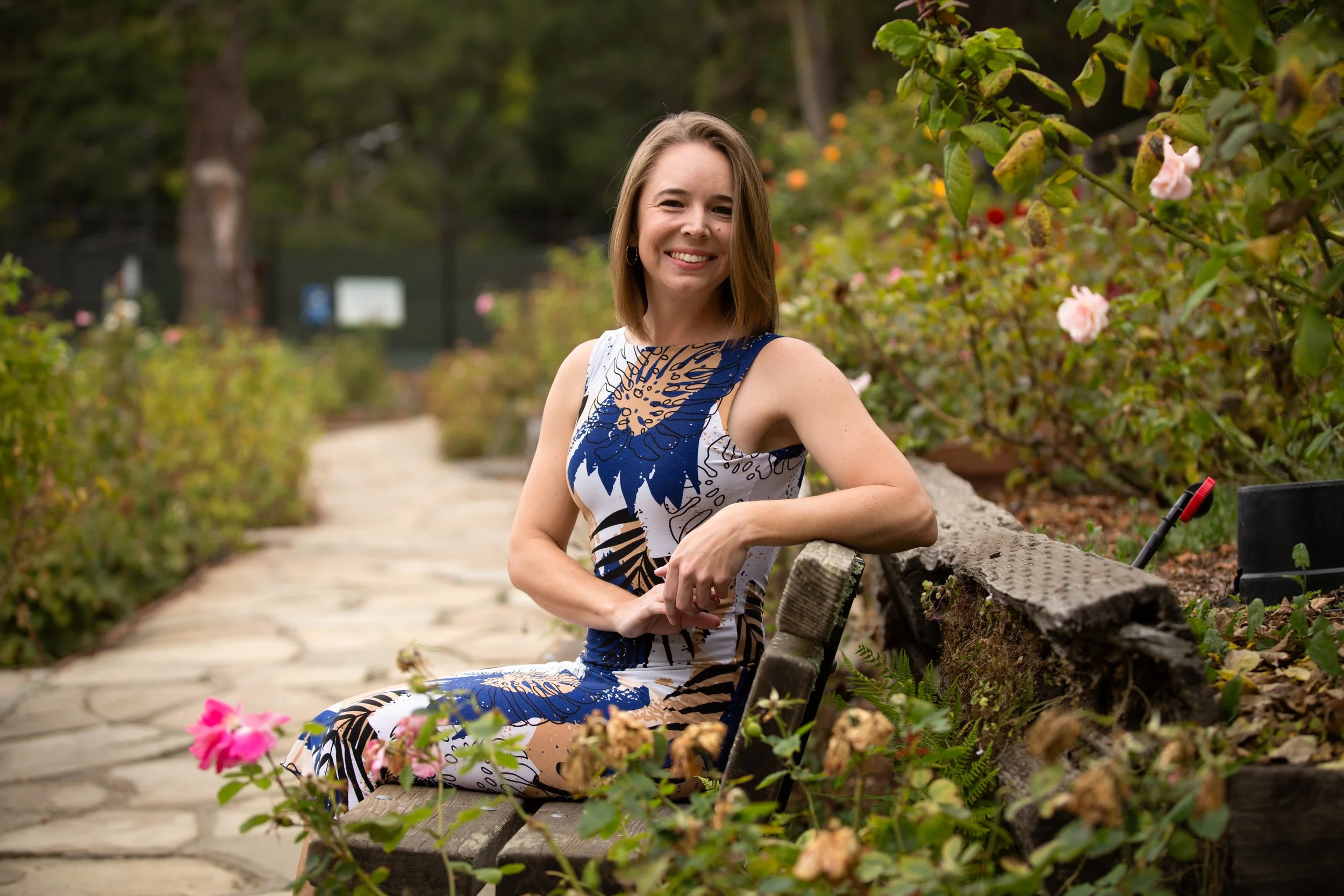 Smiling woman in a floral dress sitting on a wooden bench in a garden with pink flowers.