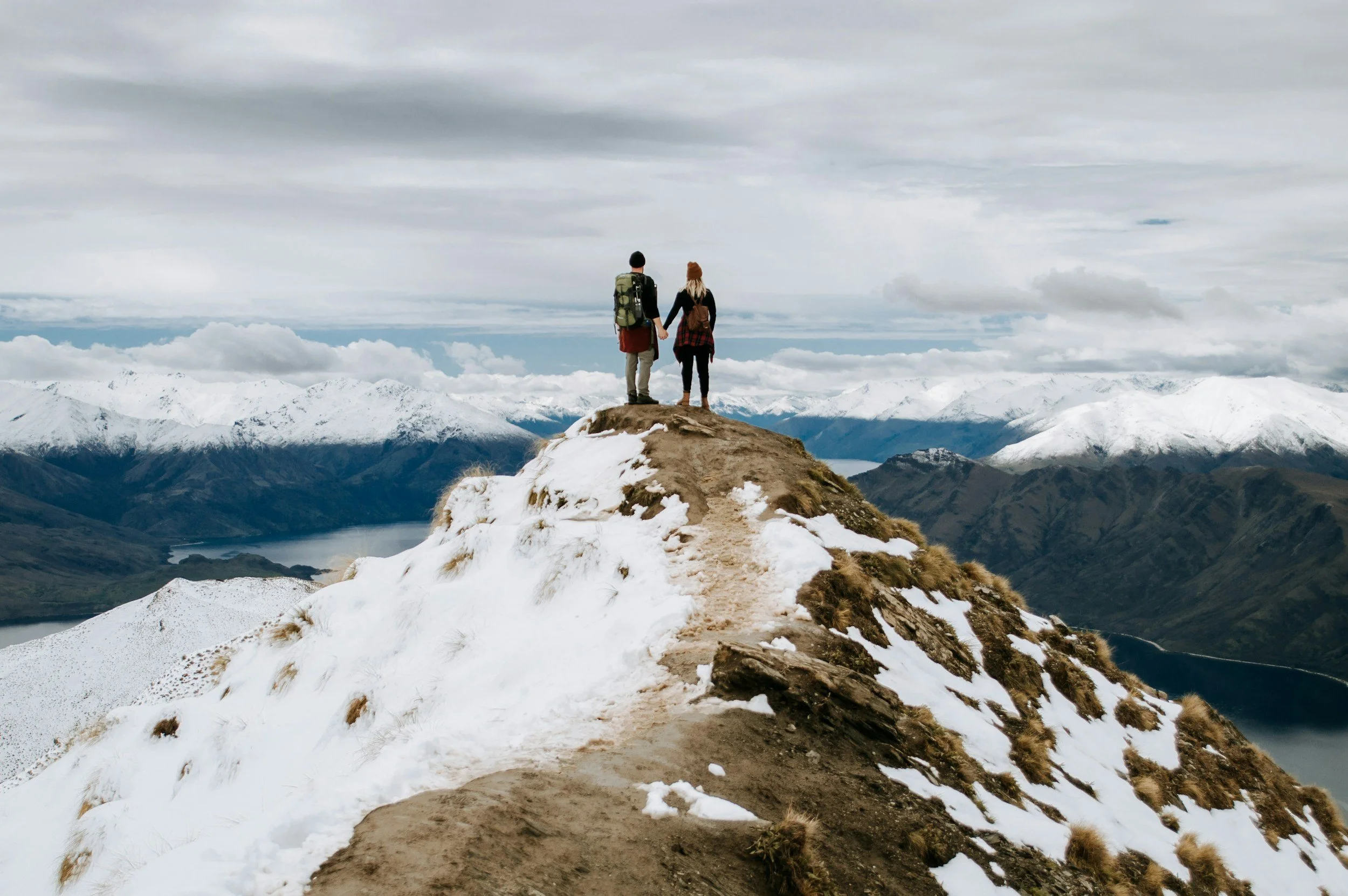 Two hikers holding hands standing on a snow-covered mountain summit overlooking a valley with lakes and snow-capped mountains in the distance.