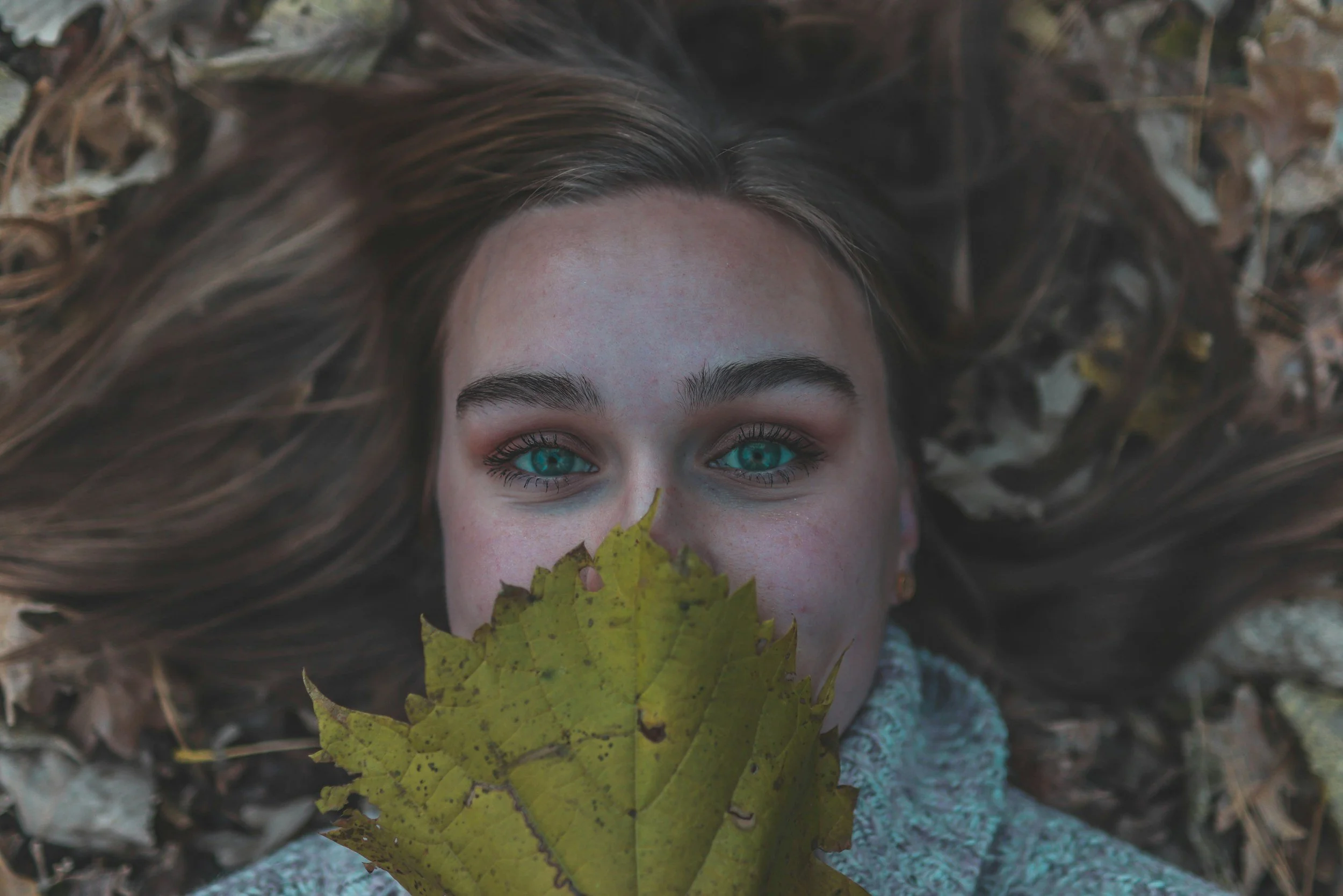 A woman lying on the ground surrounded by fallen autumn leaves, holding a yellow leaf in front of her face, with bright blue eyes visible.