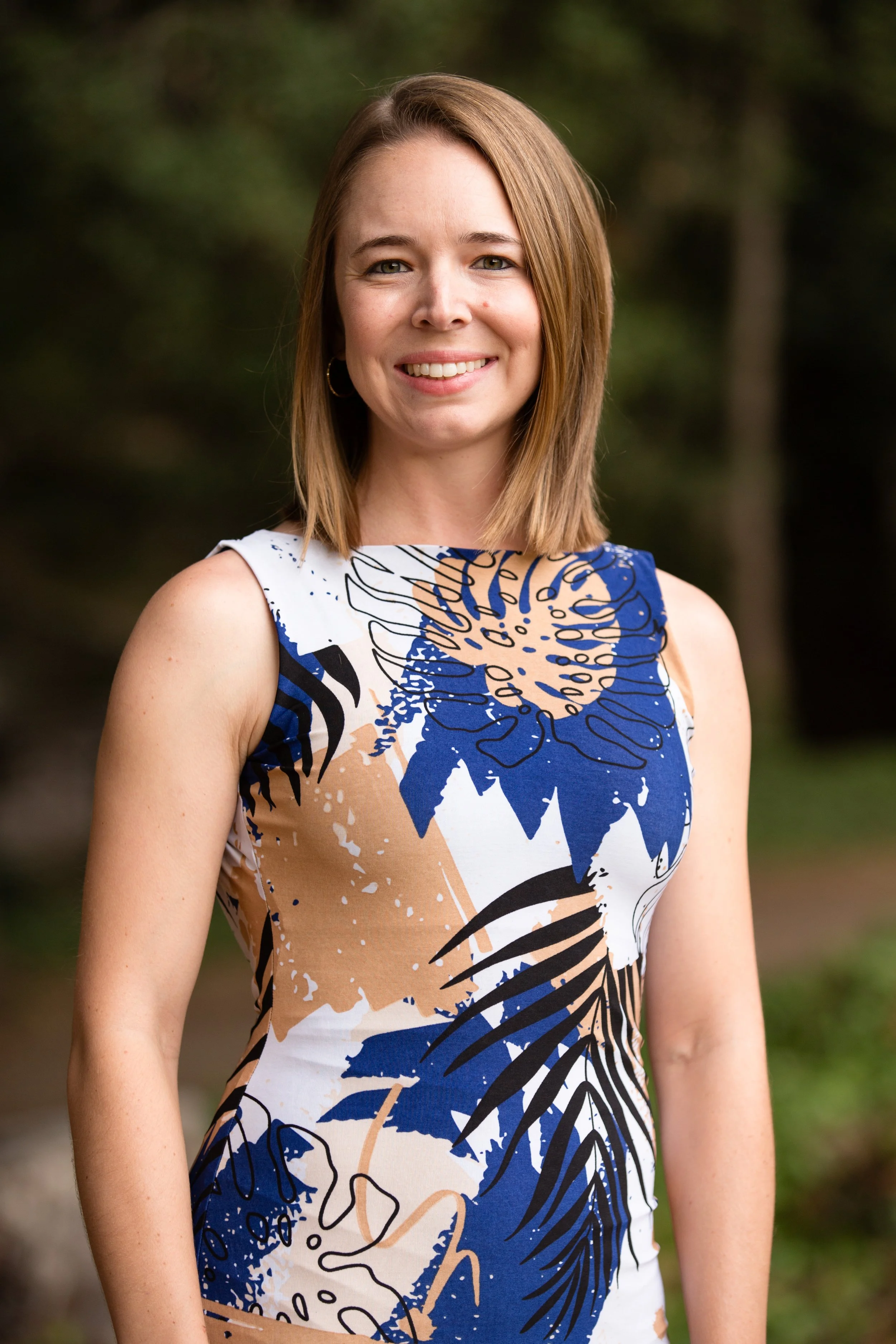 A woman with shoulder-length light brown hair, wearing a sleeveless dress with a blue, beige, black, and white abstract floral pattern, standing outdoors with a blurred green background.