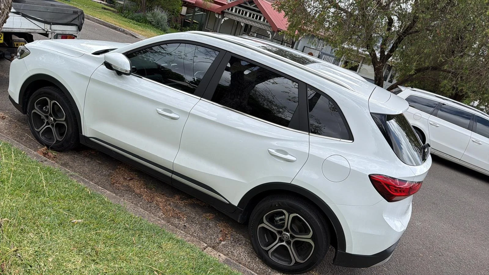 A white hatchback car parked on the side of a street with other parked vehicles nearby and trees in the background.