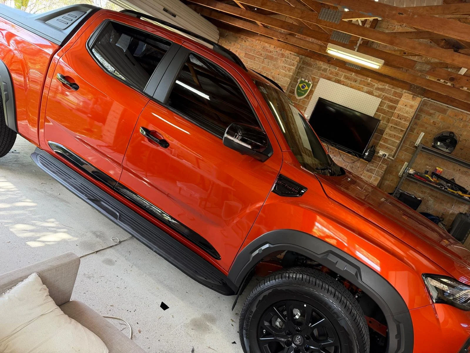 Orange pickup truck parked inside a garage with exposed brick walls and wooden ceiling. Garage has a TV mounted on the wall, a shelf with tools, and a partially visible beige couch in the foreground.
