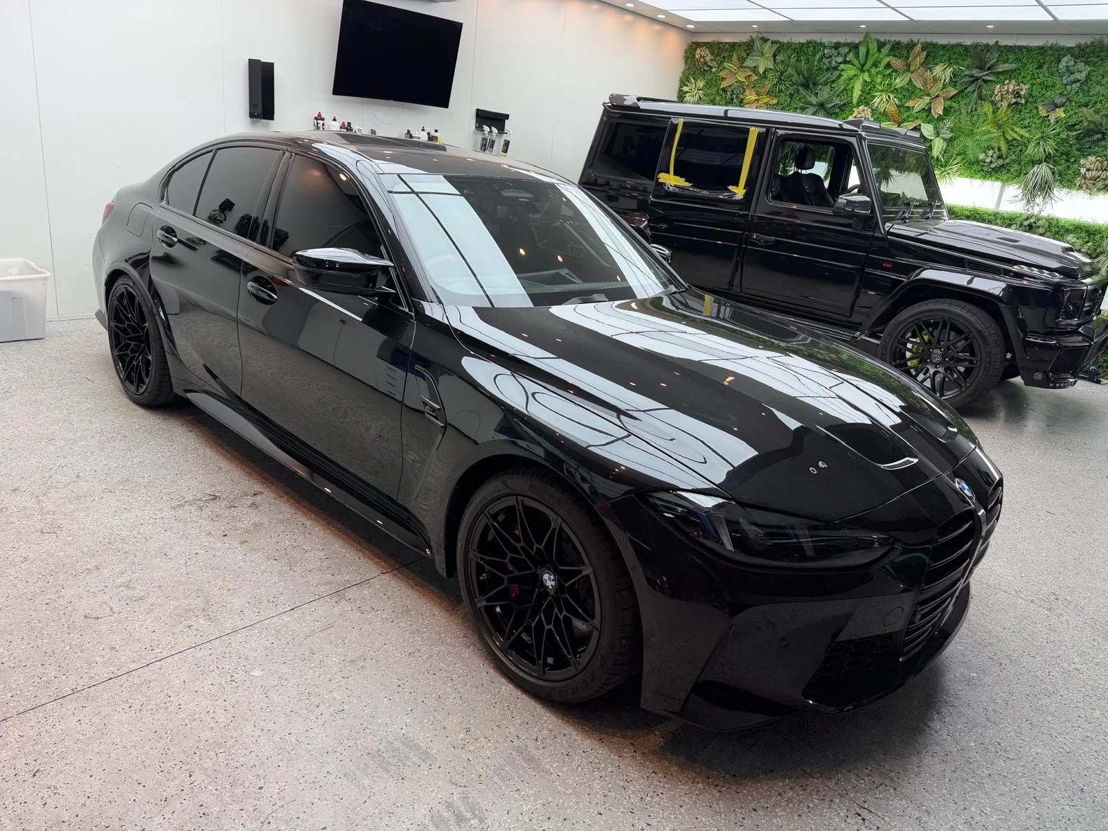 A black BMW car parked indoors next to a black luxury SUV, with a green wall and a large TV in the background.
