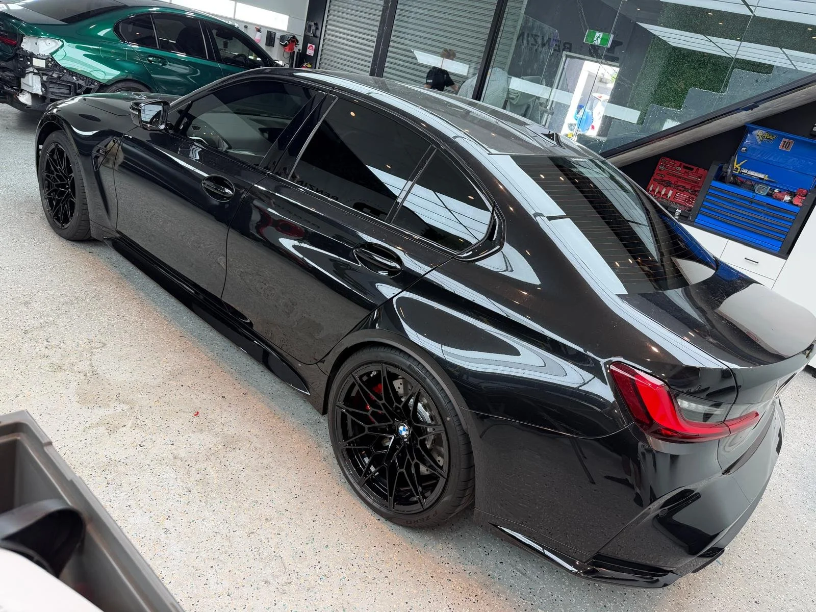 A black BMW sedan with black aftermarket wheels parked inside a car dealership or garage, with some other cars and tools in the background.