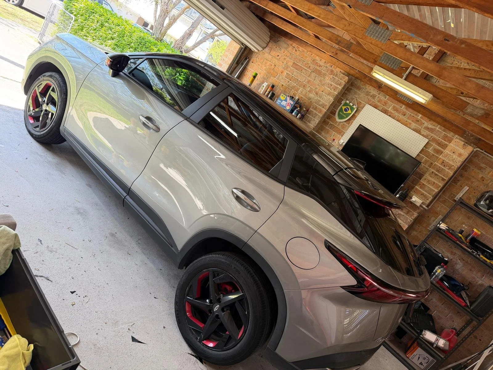 A silver and black Tesla Model Y parked inside a garage with a brick wall and wooden ceiling. The garage has shelves with tools and items, a TV, and a wall-mounted flag of Brazil.