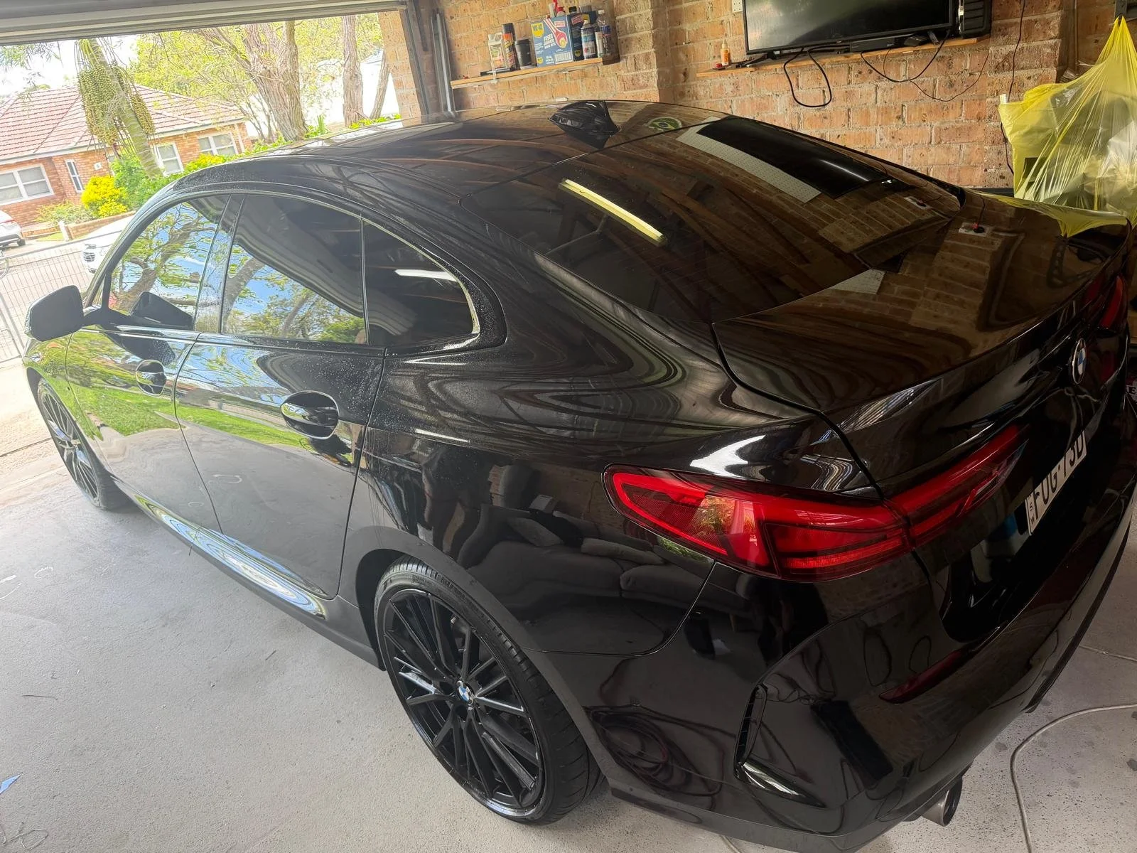 Black sedan car parked inside a garage with brick walls, partially covered with a yellow plastic sheet, reflecting trees and the sky through a window.