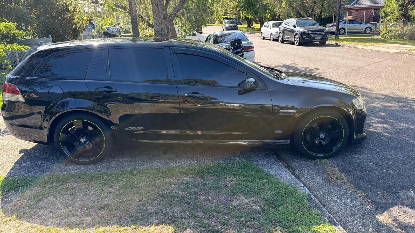 Black car parked in a driveway with a grassy area and trees in the background, with other cars parked on the street.