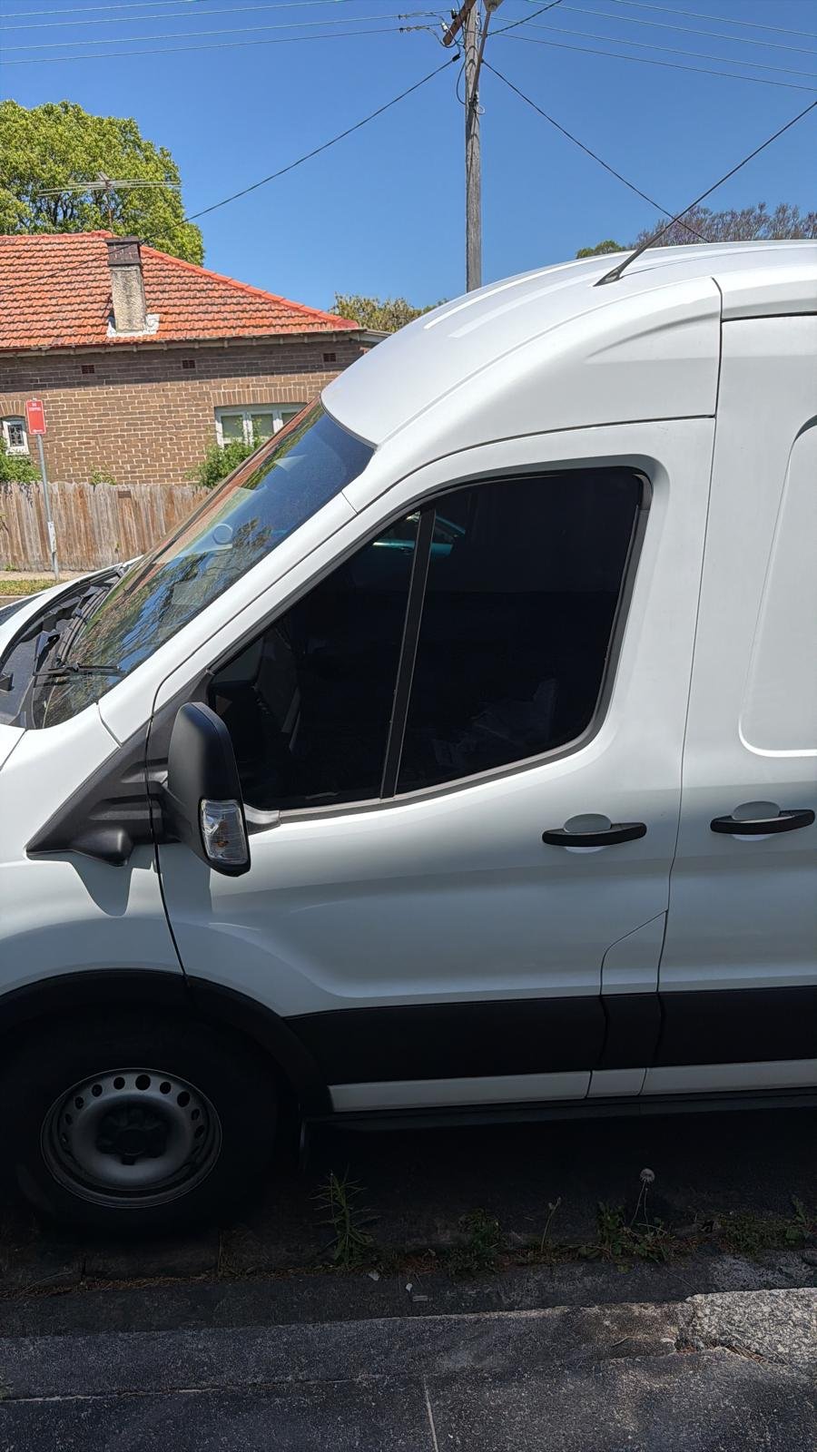 Side view of a white commercial van parked on the street with a house in the background, a blue sky, and electrical wires overhead.