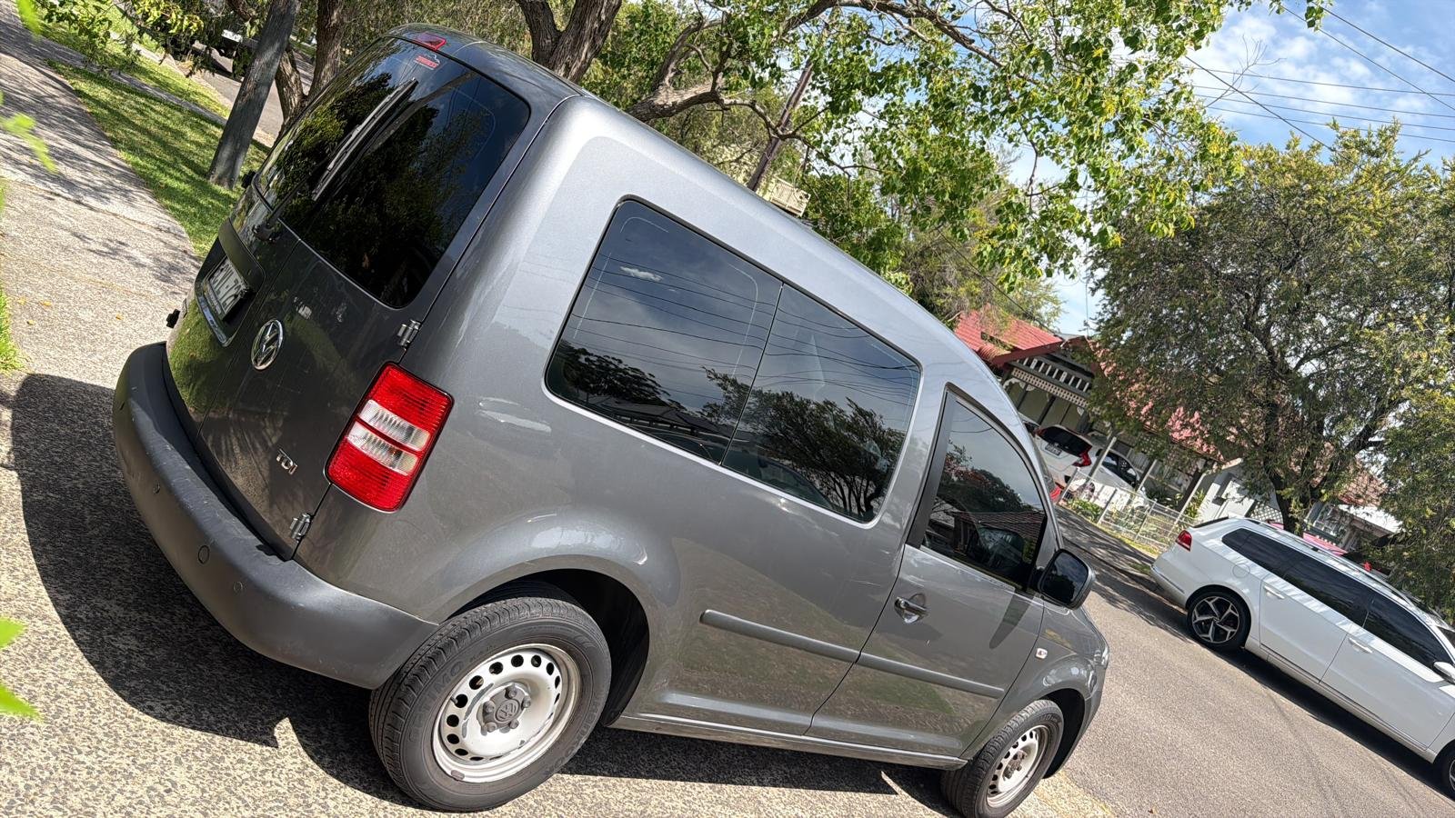 A gray Volkswagen Caddy van parked on a driveway with trees and houses in the background.