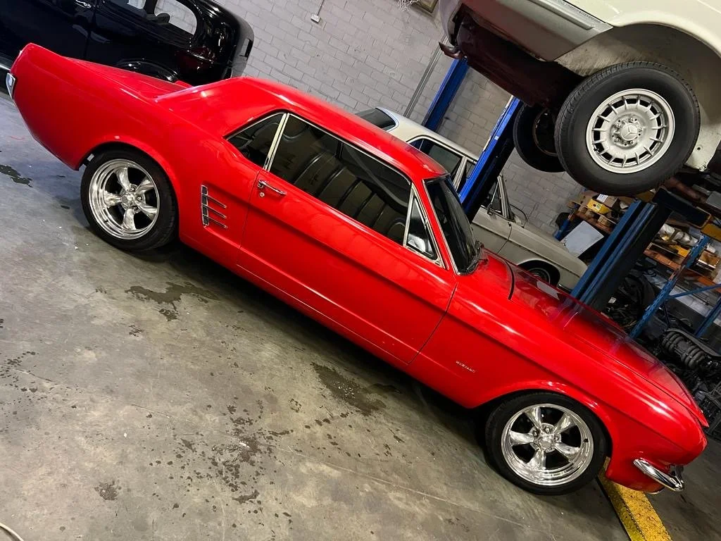 Red vintage Corvette sports car with chrome wheels, parked indoors in a garage with other cars and tools visible.