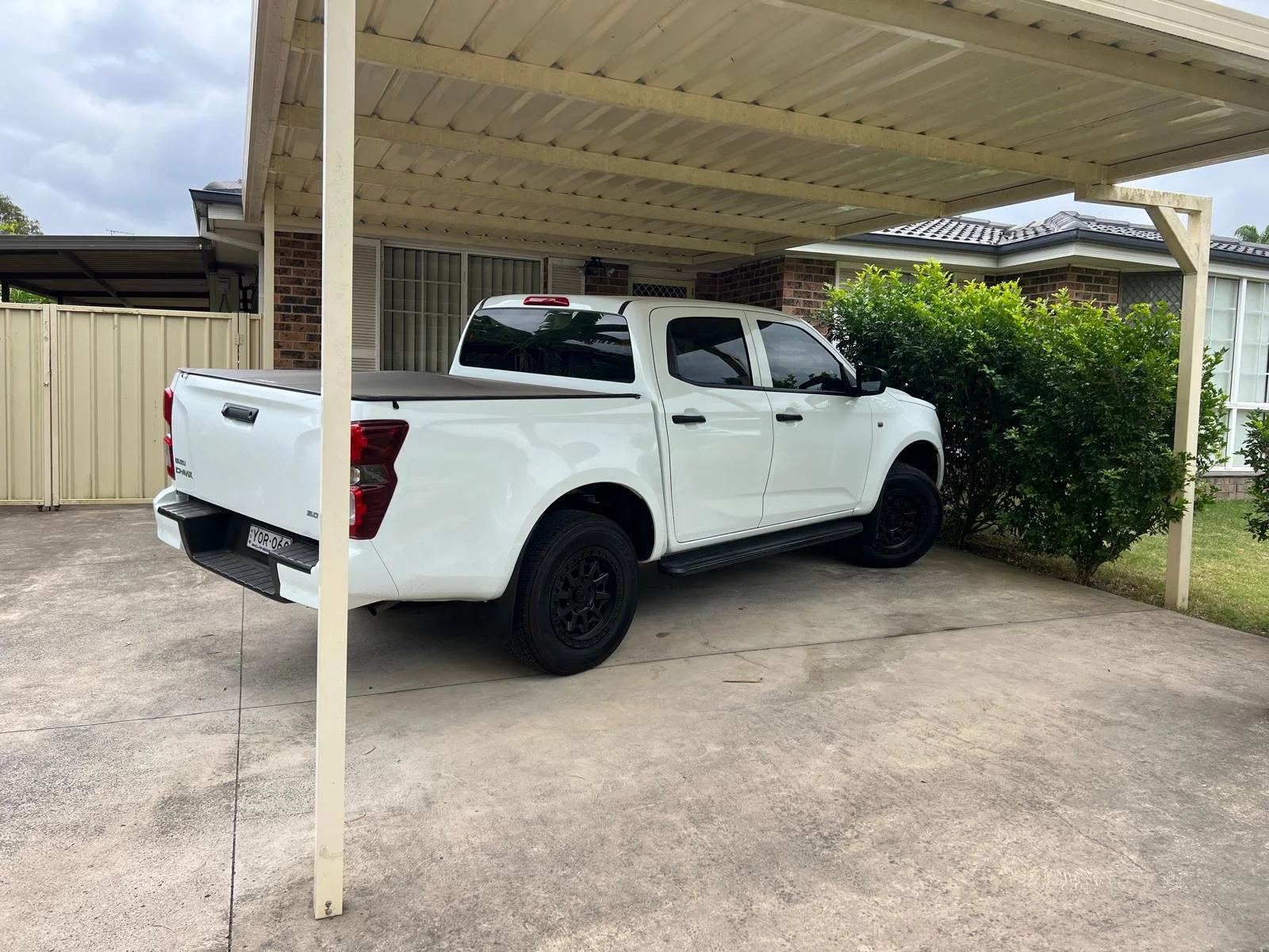 White pickup truck parked under a carport in a residential driveway, with bushes and a house in the background.