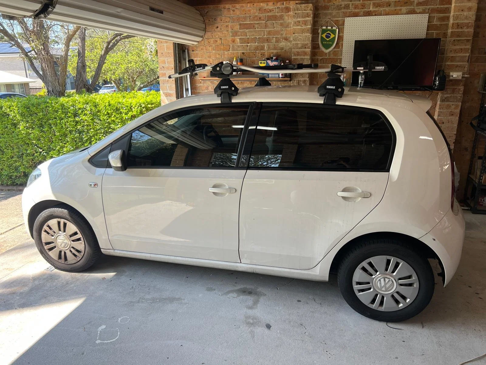 A white Volkswagen compact car parked inside a garage with a brick wall. The car has a roof rack with a ski or snowboard carrier. Outside the garage, trees and parked cars are visible.