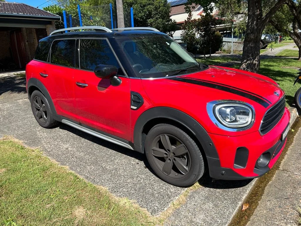 Red MINI Cooper with black racing stripes parked on a residential street with trees and houses in the background.