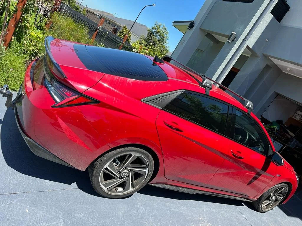 Red electric vehicle parked in driveway with solar panels on the roof, next to a house and garden.