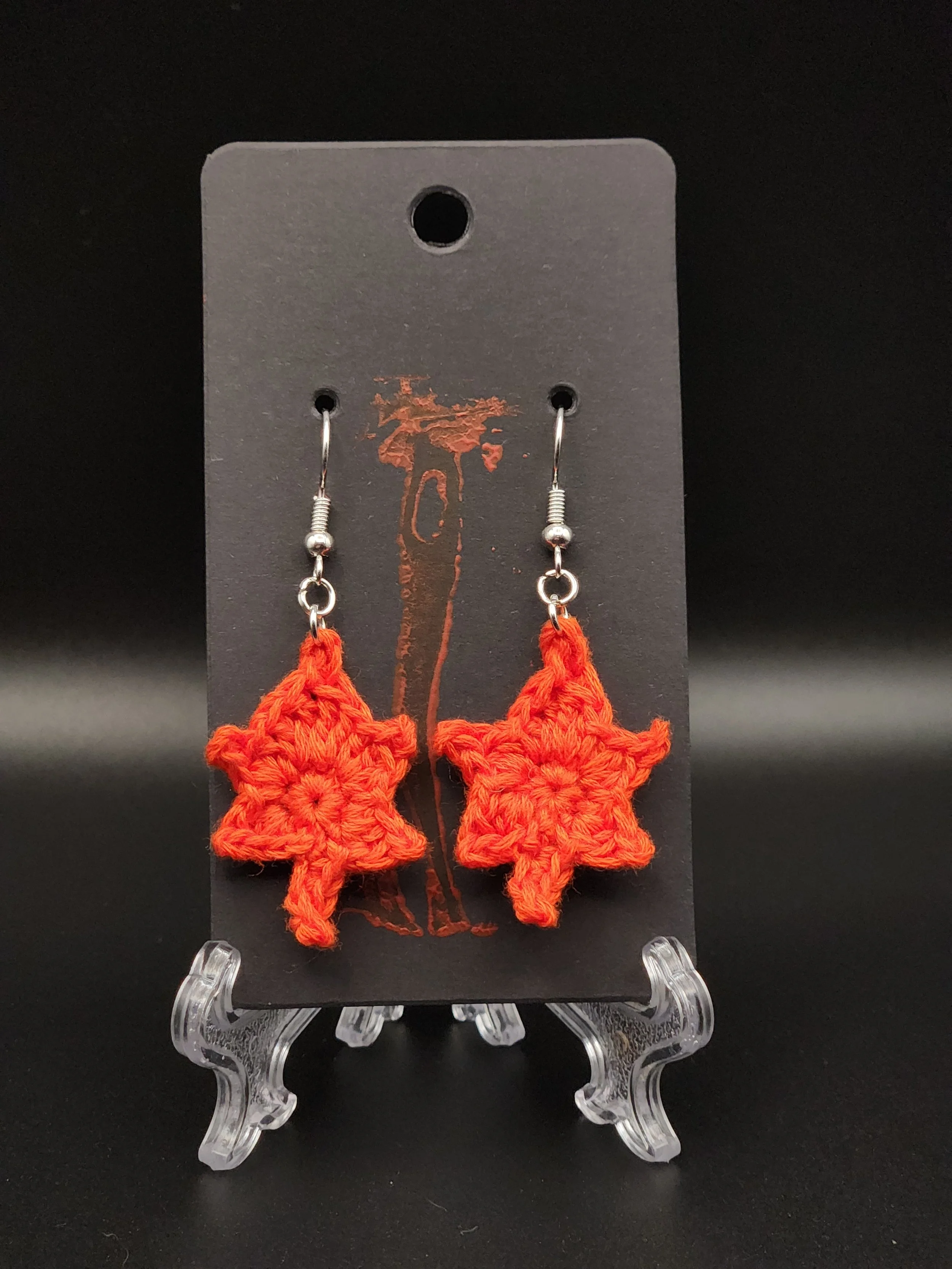 Orange crochet earrings shaped like Christmas trees hanging on a black display card.