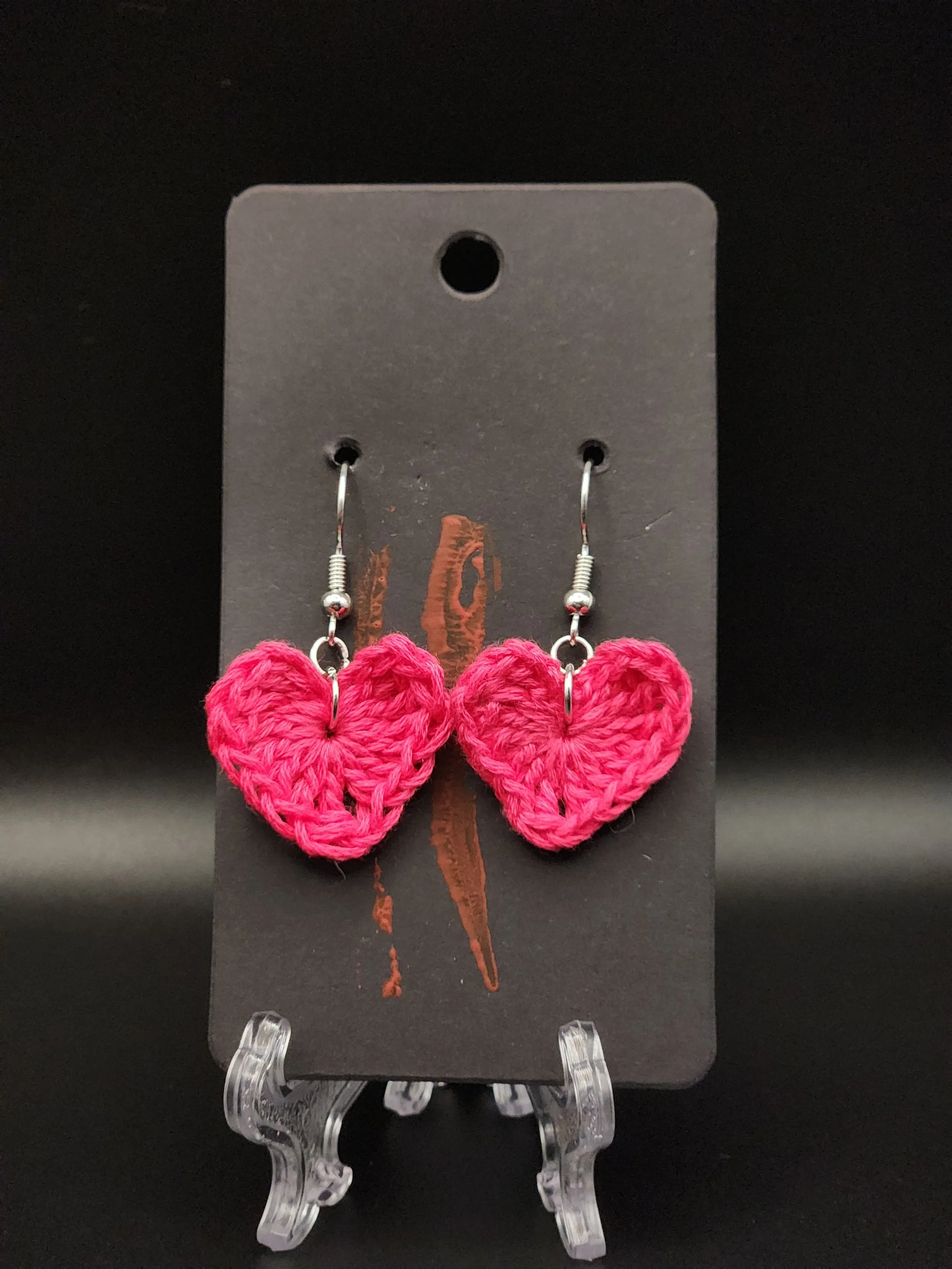 Pink crocheted heart-shaped earrings displayed on a black card with a red lightning bolt design, mounted on a clear plastic stand.