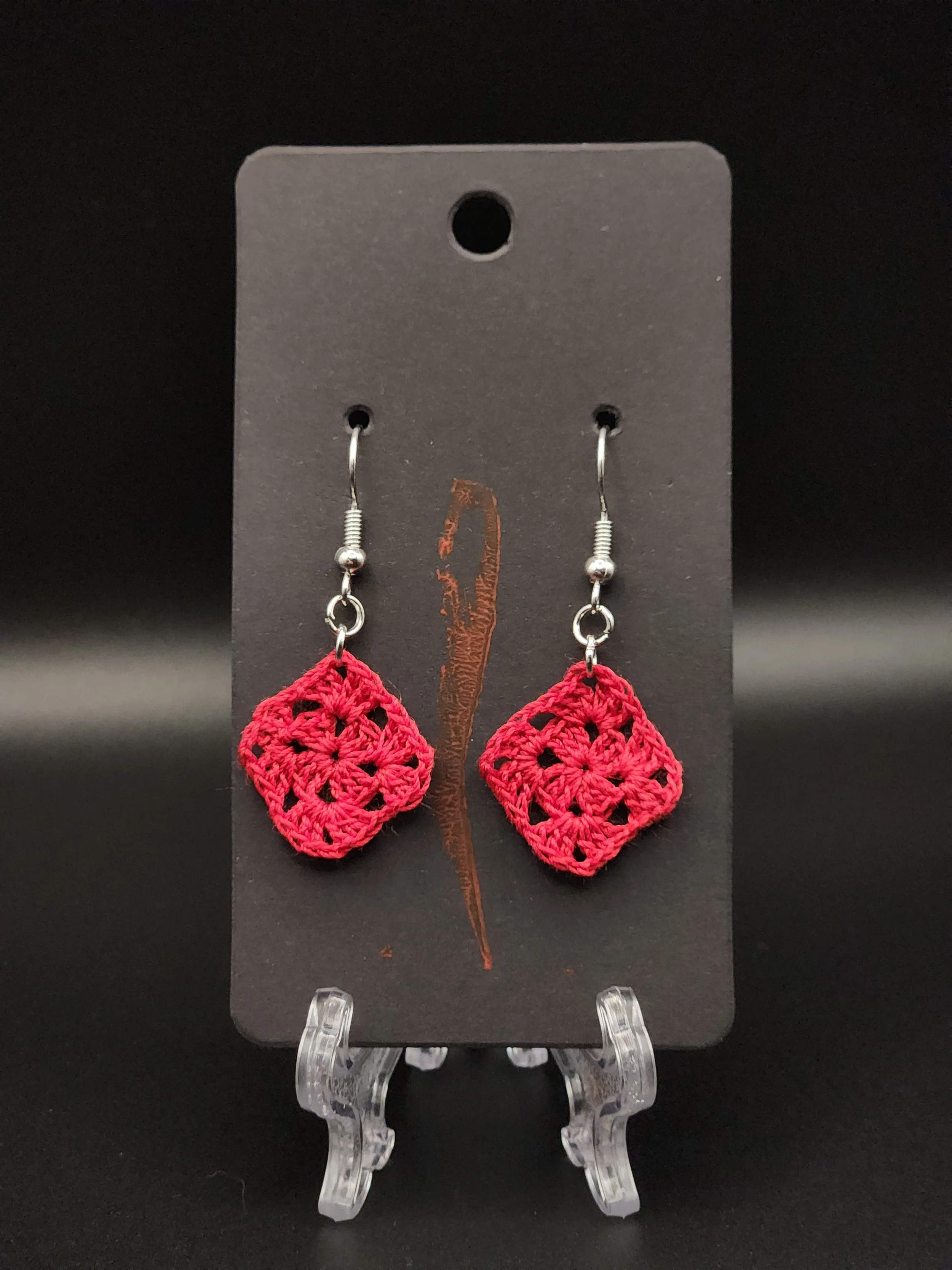 Red crocheted square earrings hanging on a black display card.
