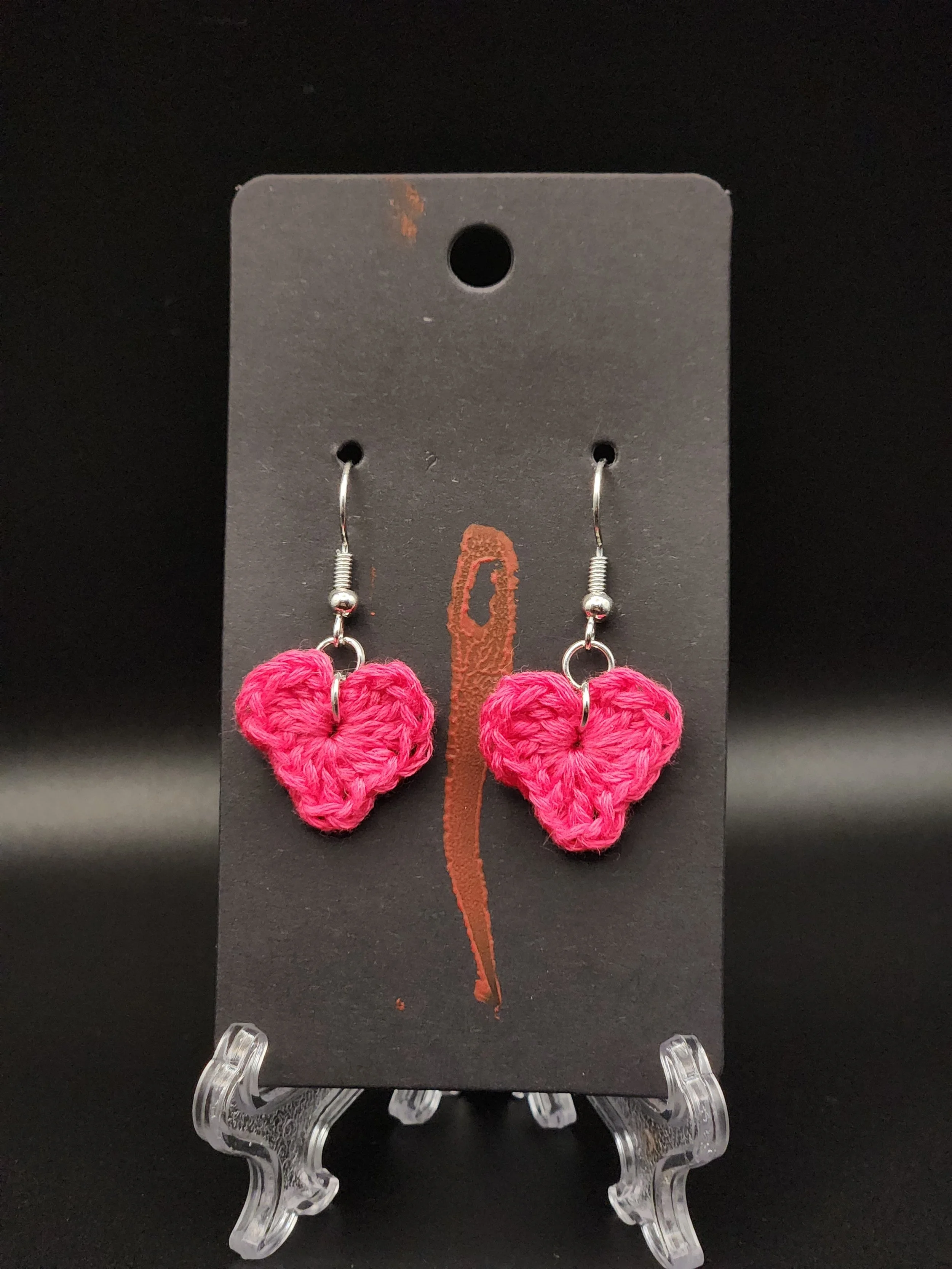 Pink crochet heart earrings displayed on a black cardboard holder with a red chili pepper graphic, against a black background.
