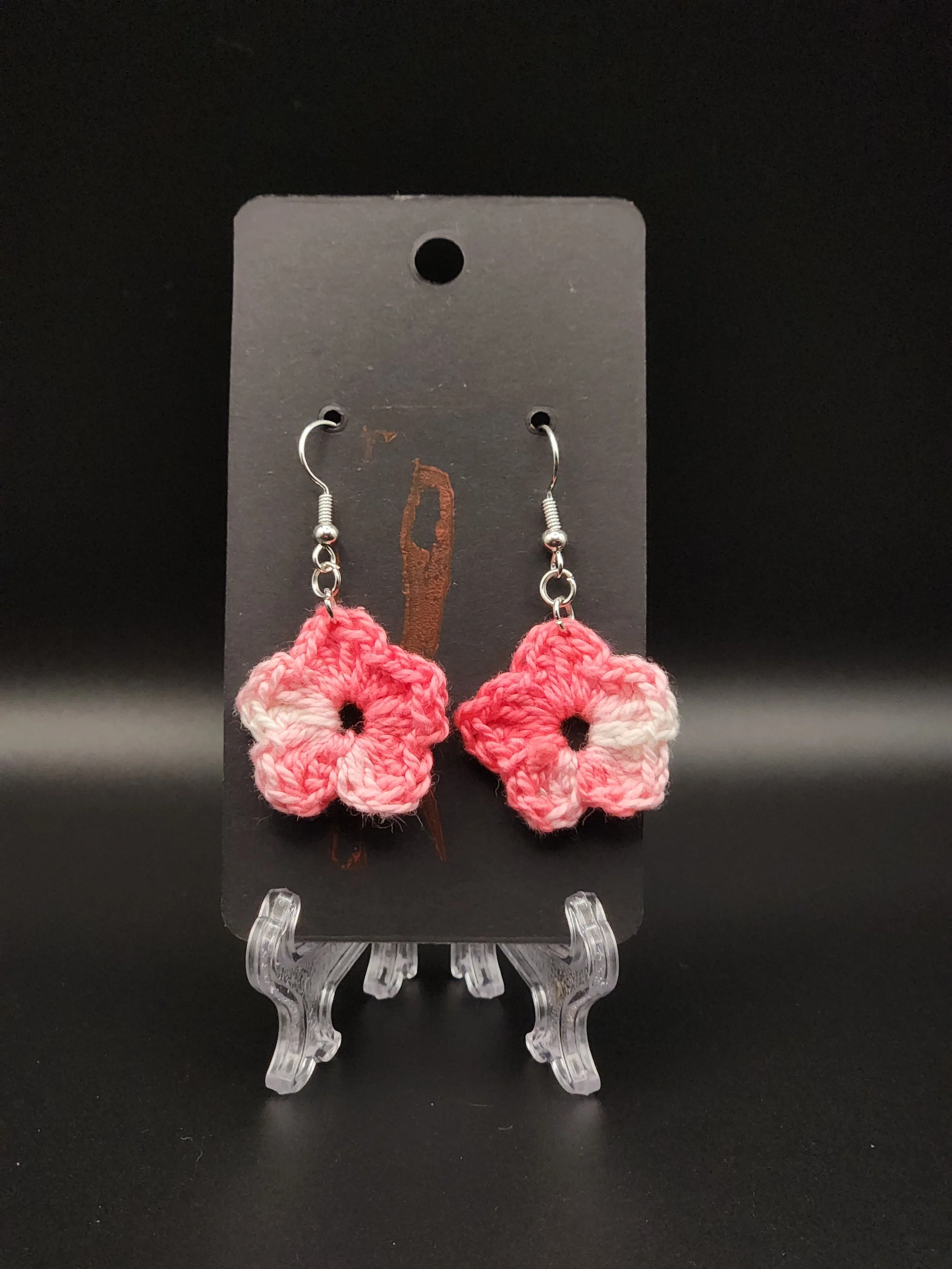 Pink and white crocheted flower earrings displayed on a black card background.