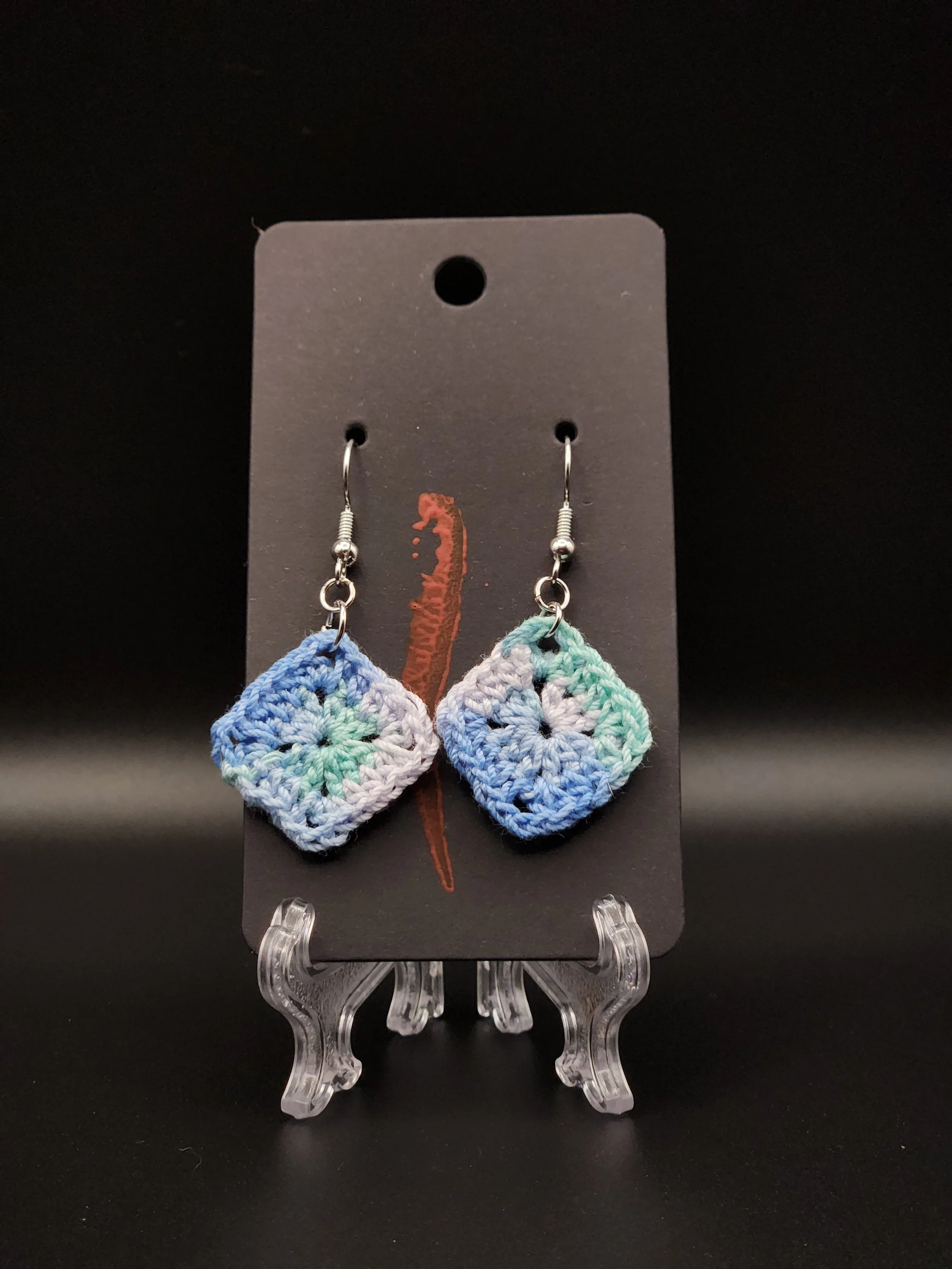 Colorful crochet earrings with square patterns on display on a black card.