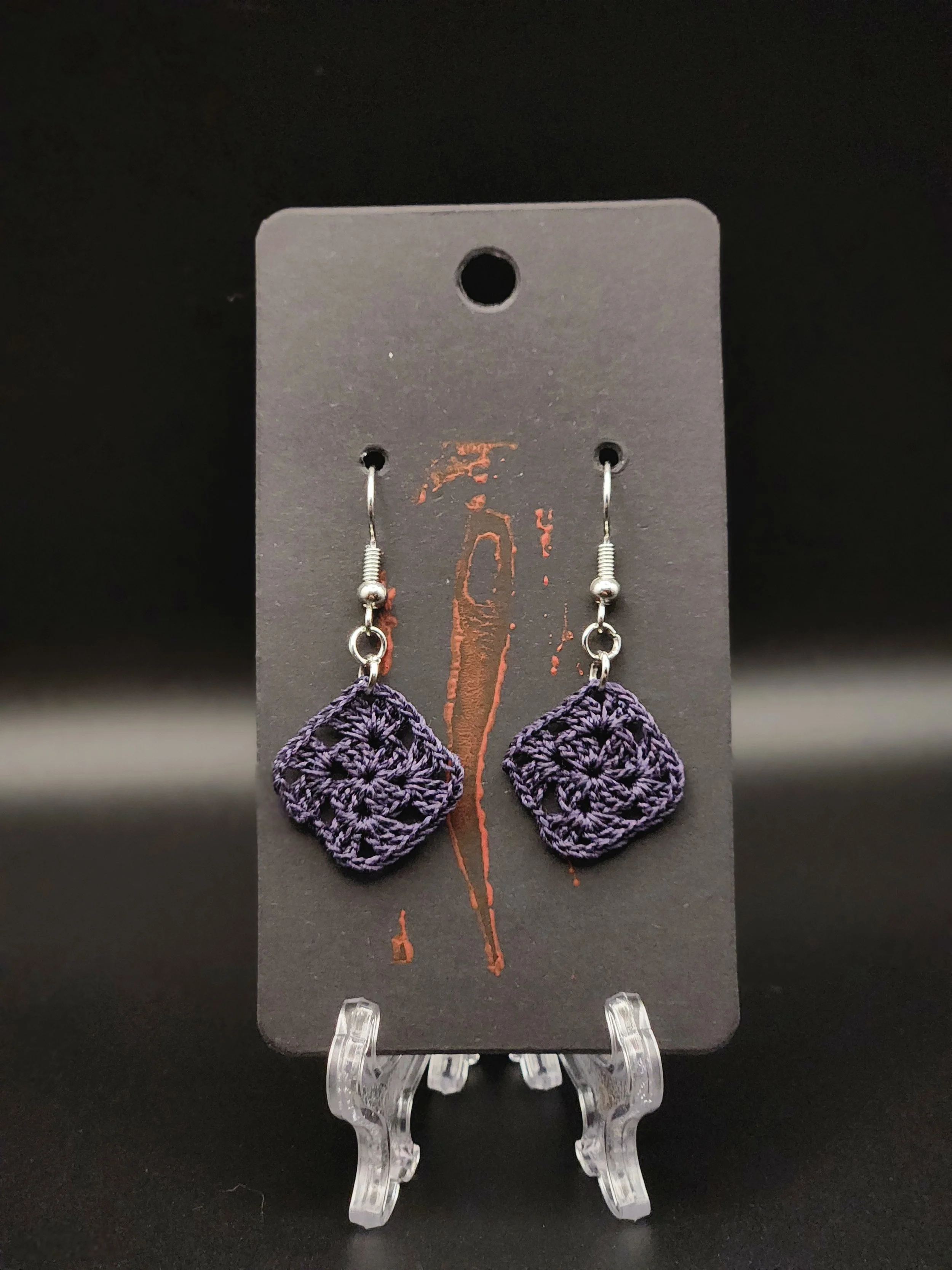 Pair of purple crocheted earrings hanging on a black display card with a small hole at the top and an orange design. The earrings are attached to metal hooks.