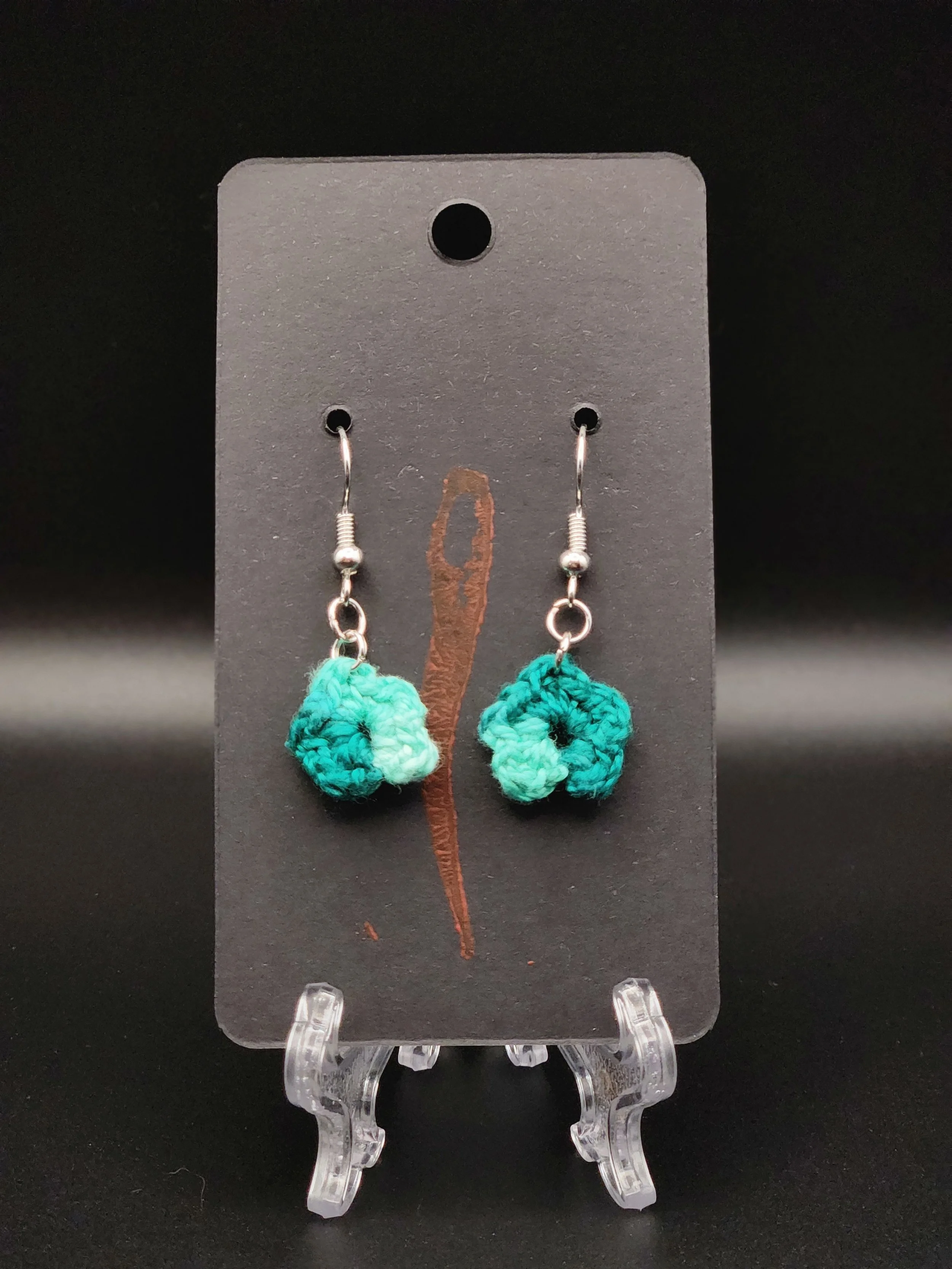 Colorful crochet earrings hanging on a black display card with a red and brown tear mark in the background.