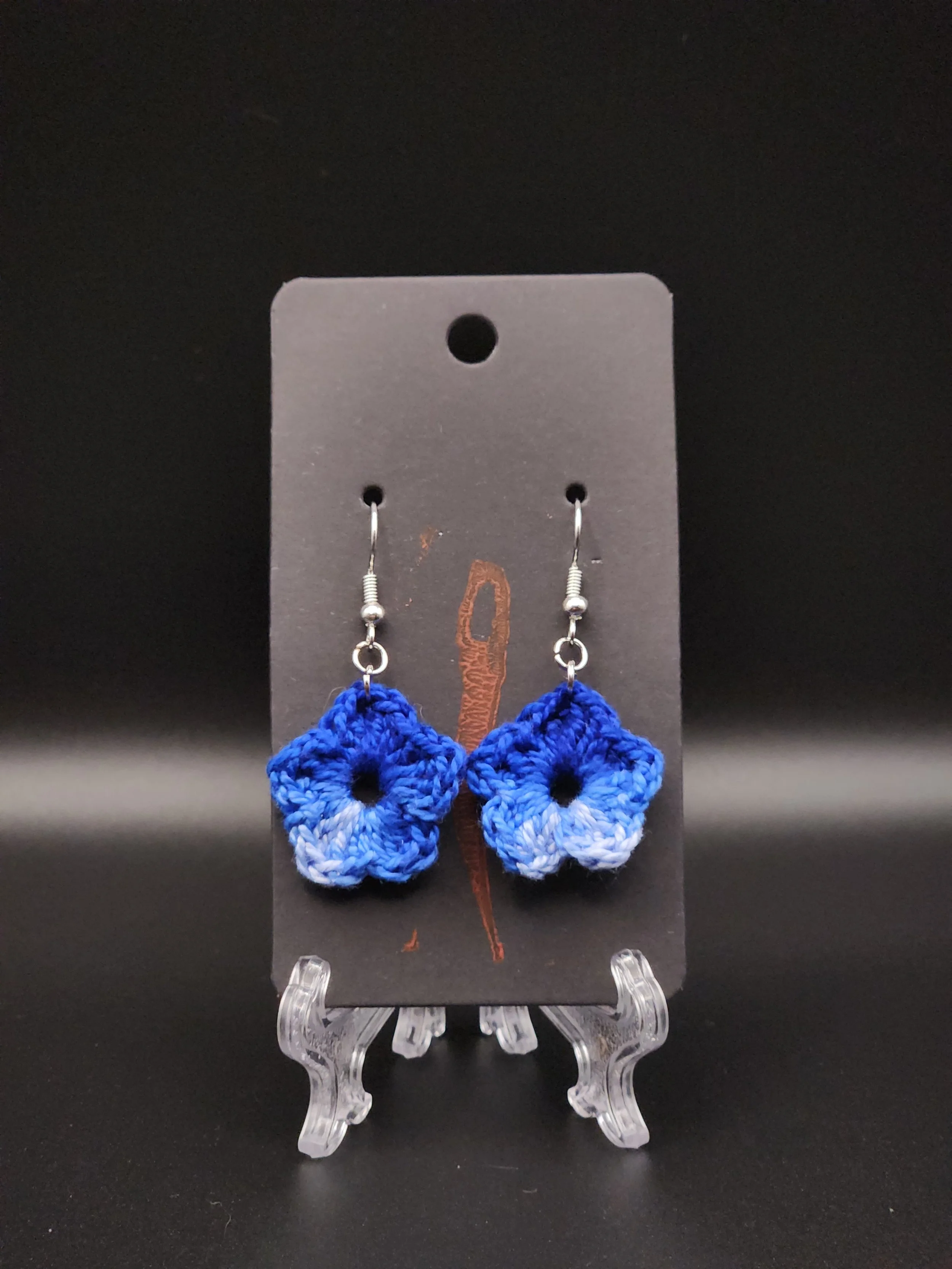 Blue crocheted flower earrings on a black jewelry display card, with a small clear plastic stand on a black surface.