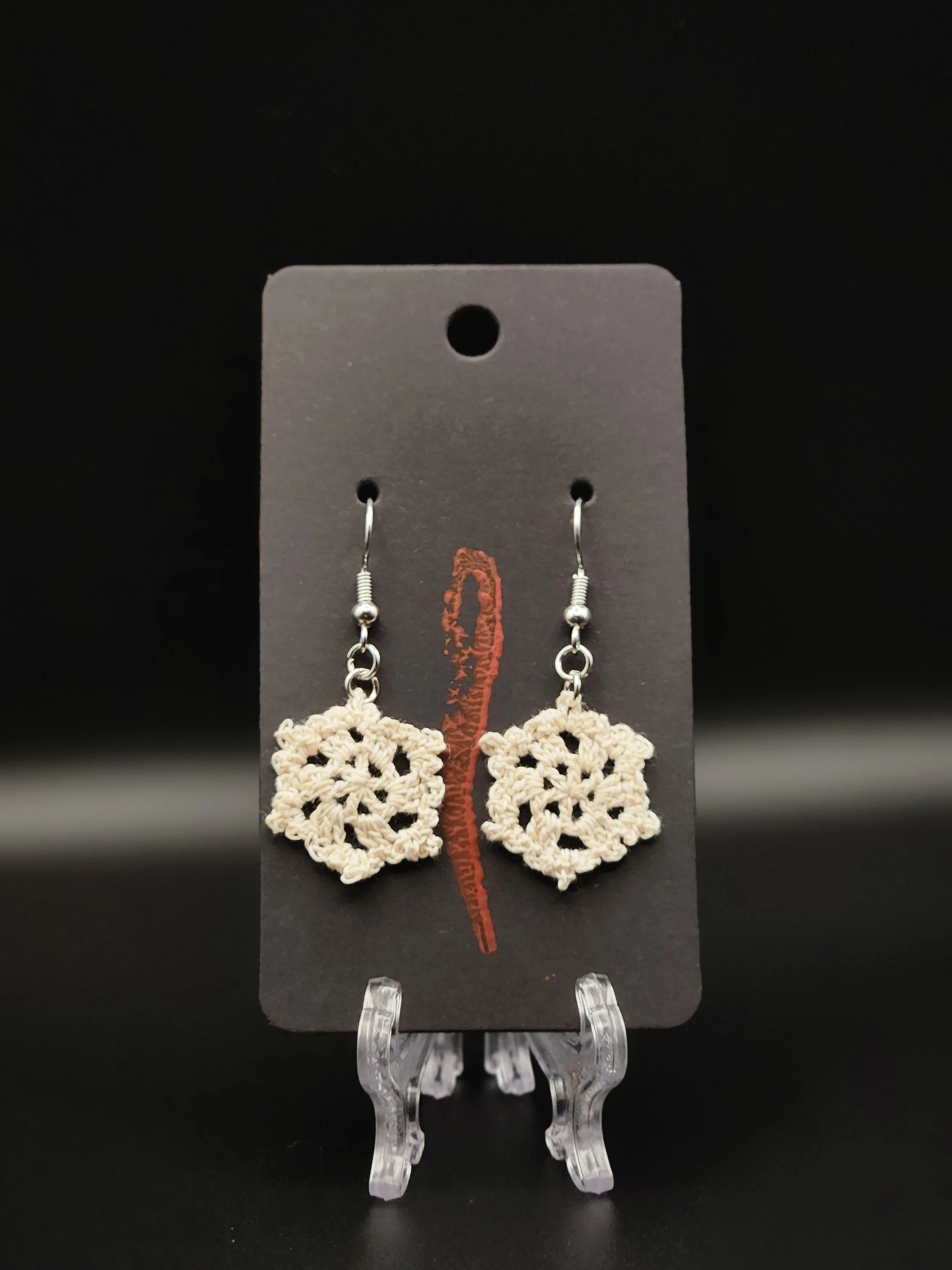 Pair of earrings with cream-colored crochet snowflake designs hanging from metal hooks on black display card.