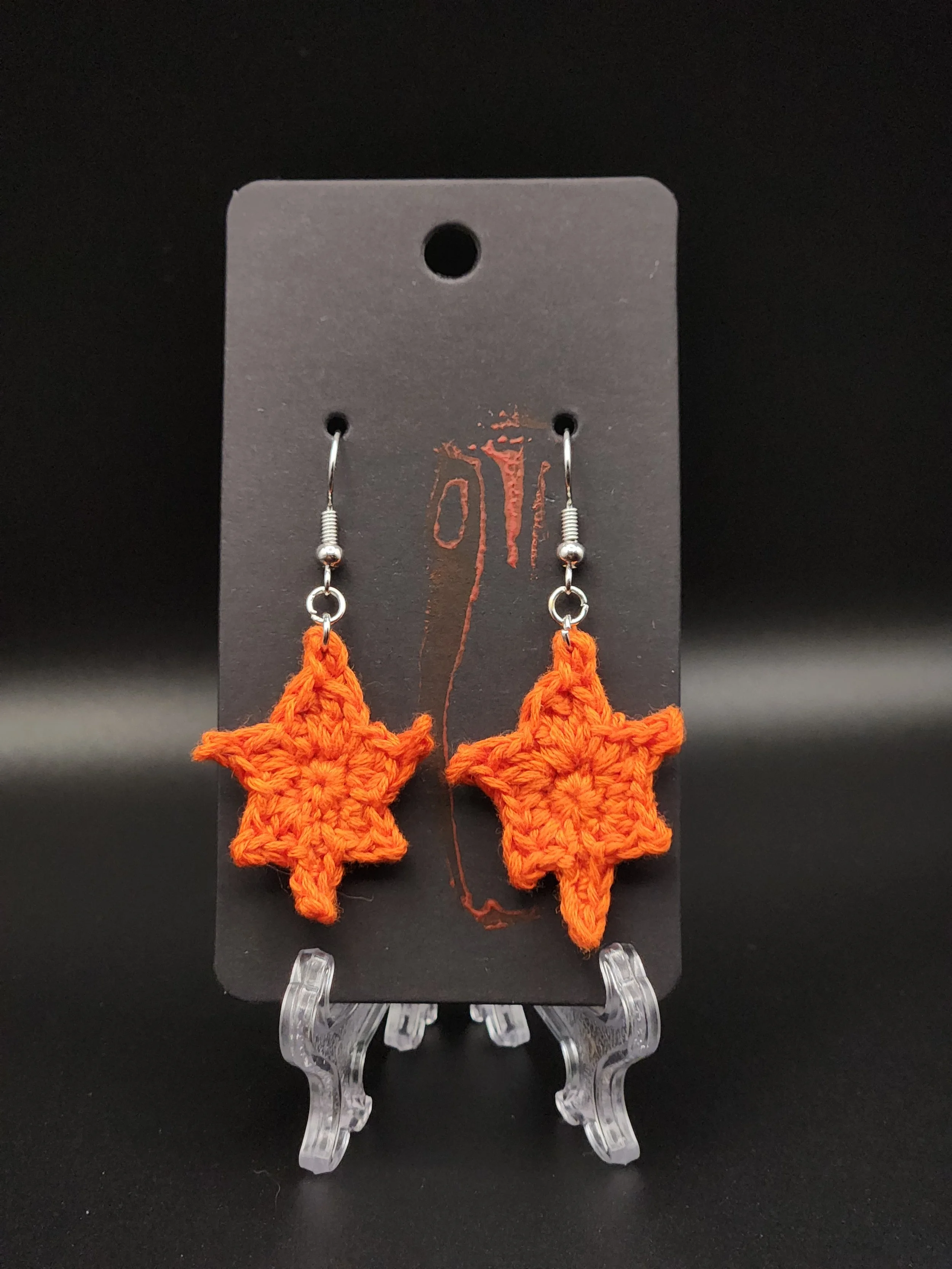 Orange crocheted star-shaped earrings on a black display card against a black background.