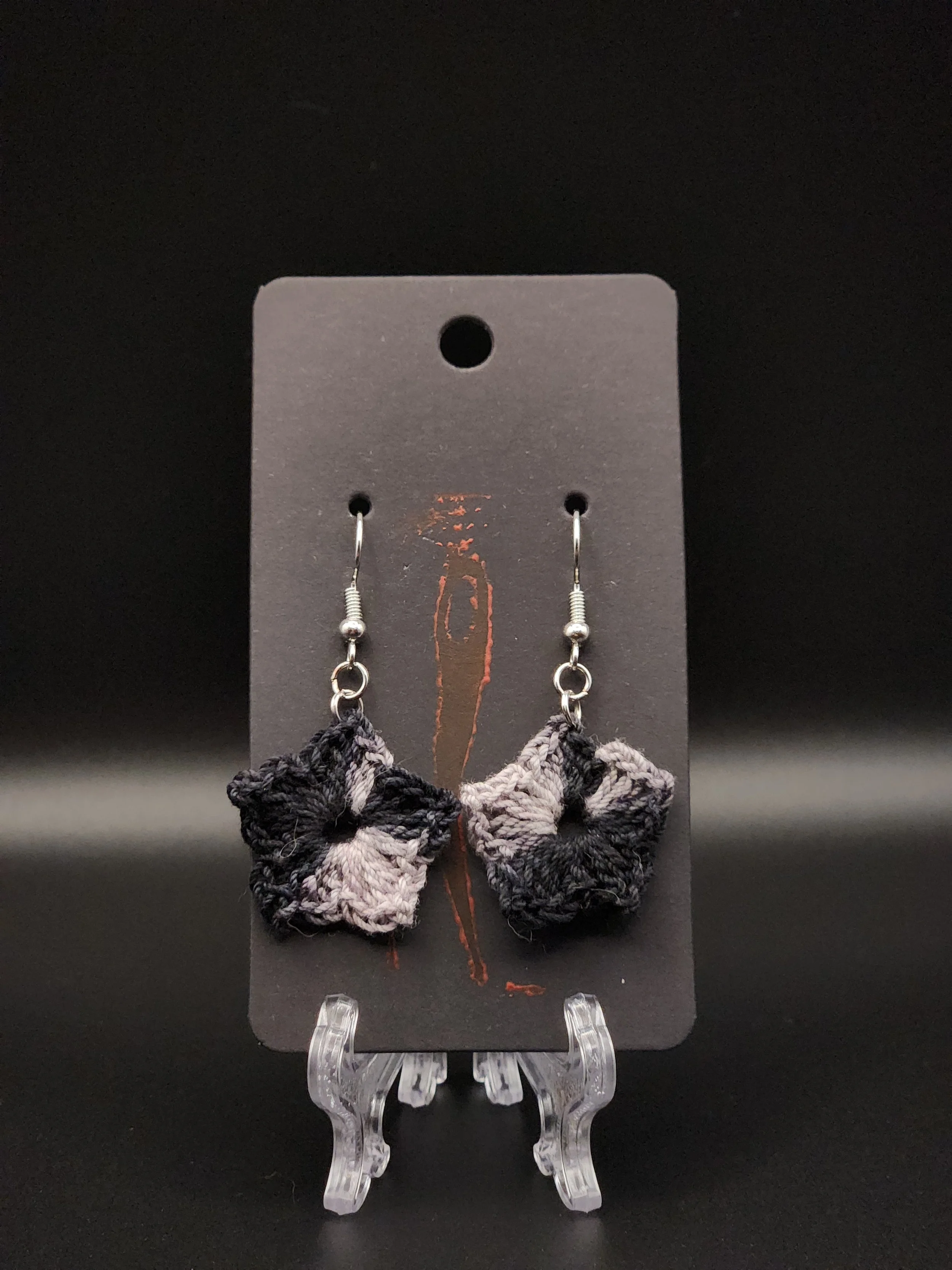 A pair of black, gray, and white fabric earrings with a satin-like finish, hanging from silver hooks, displayed on a black board with a red design, on a transparent plastic stand.