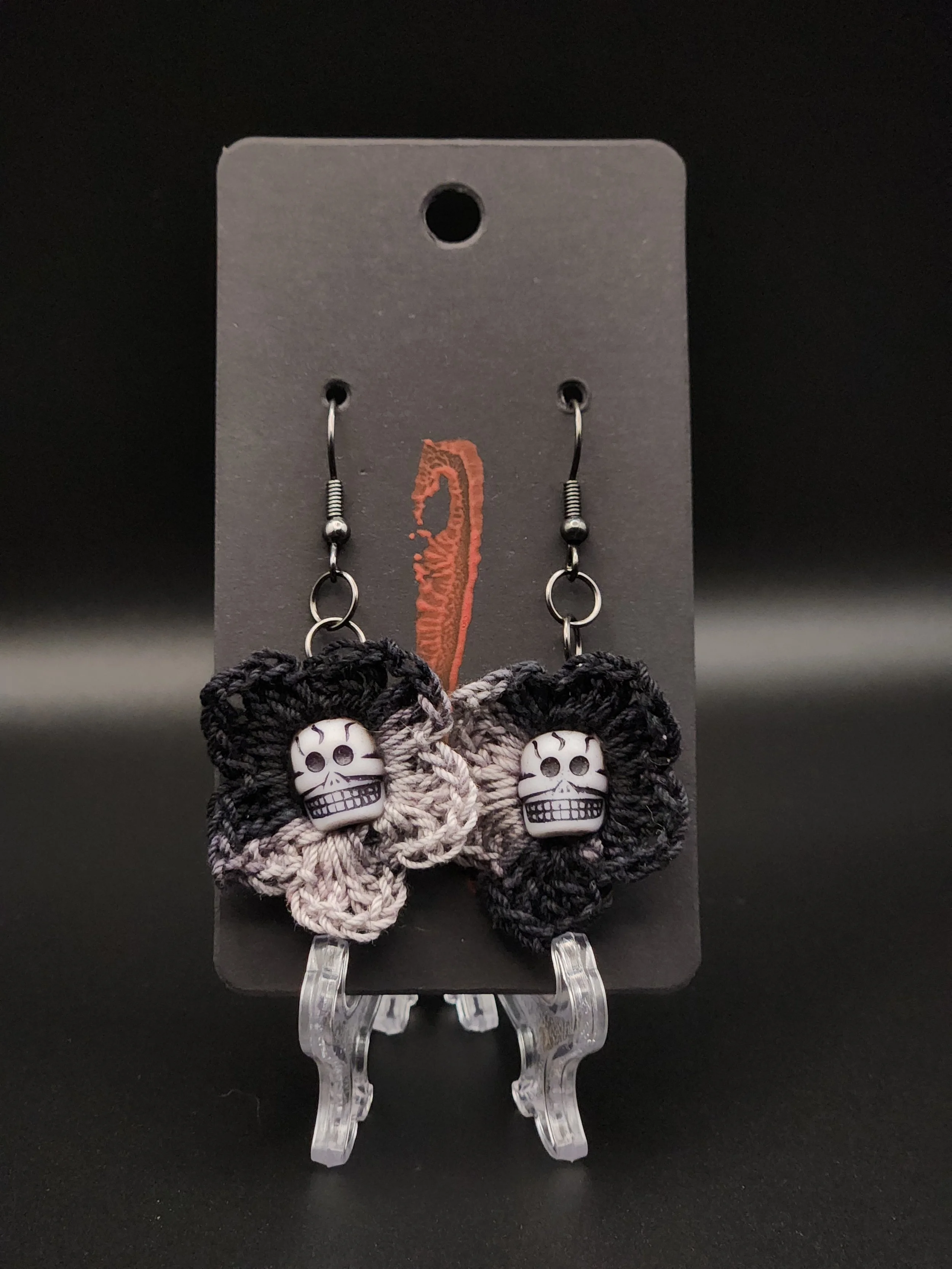 Pair of crochet earrings with skull charms and butterfly-shaped lace, displayed on a black backing card.