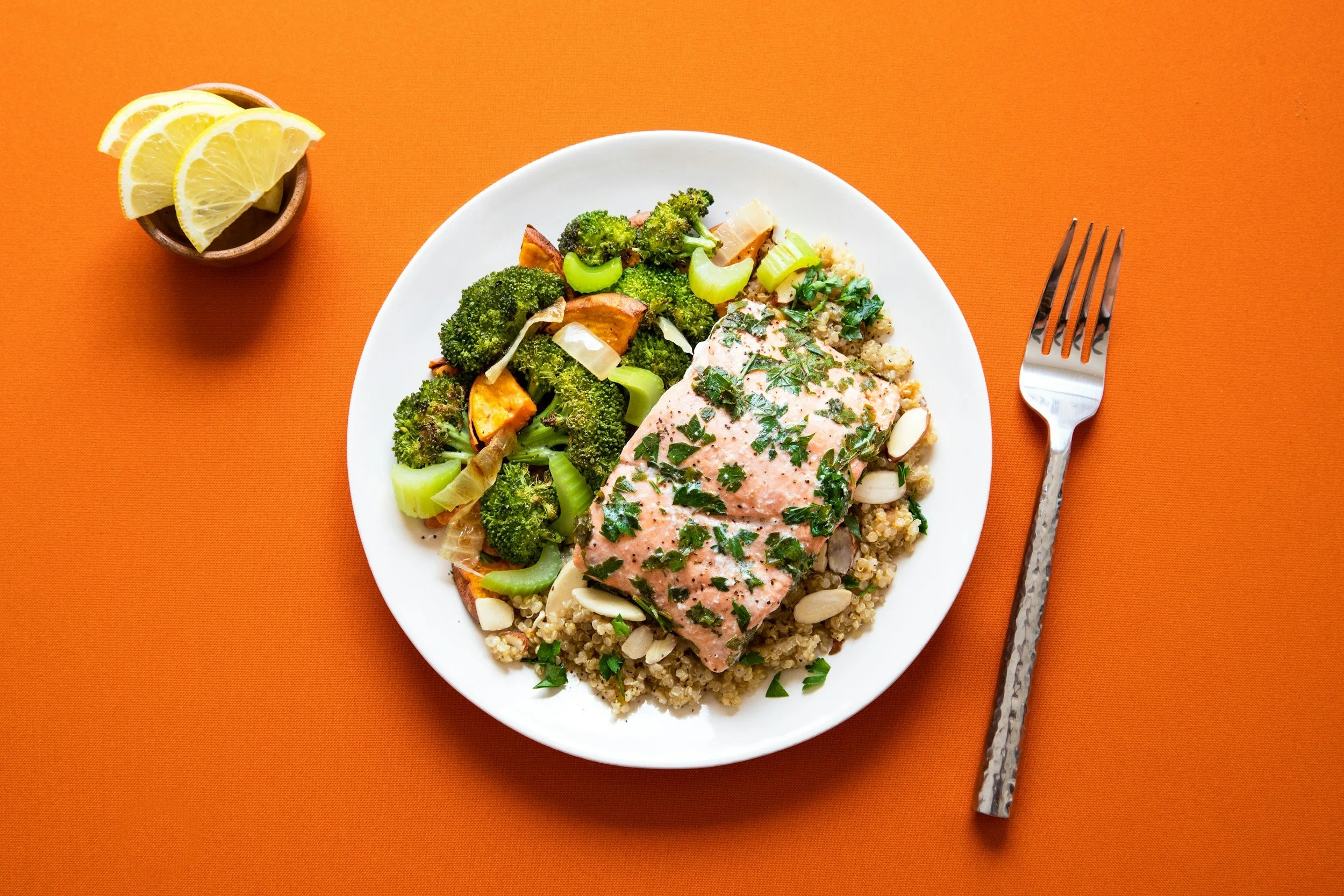 A white plate containing cooked quinoa, a piece of salmon topped with chopped herbs, and roasted broccoli and sweet potatoes, on an orange background with a fork, and a bowl of lemon wedges nearby.