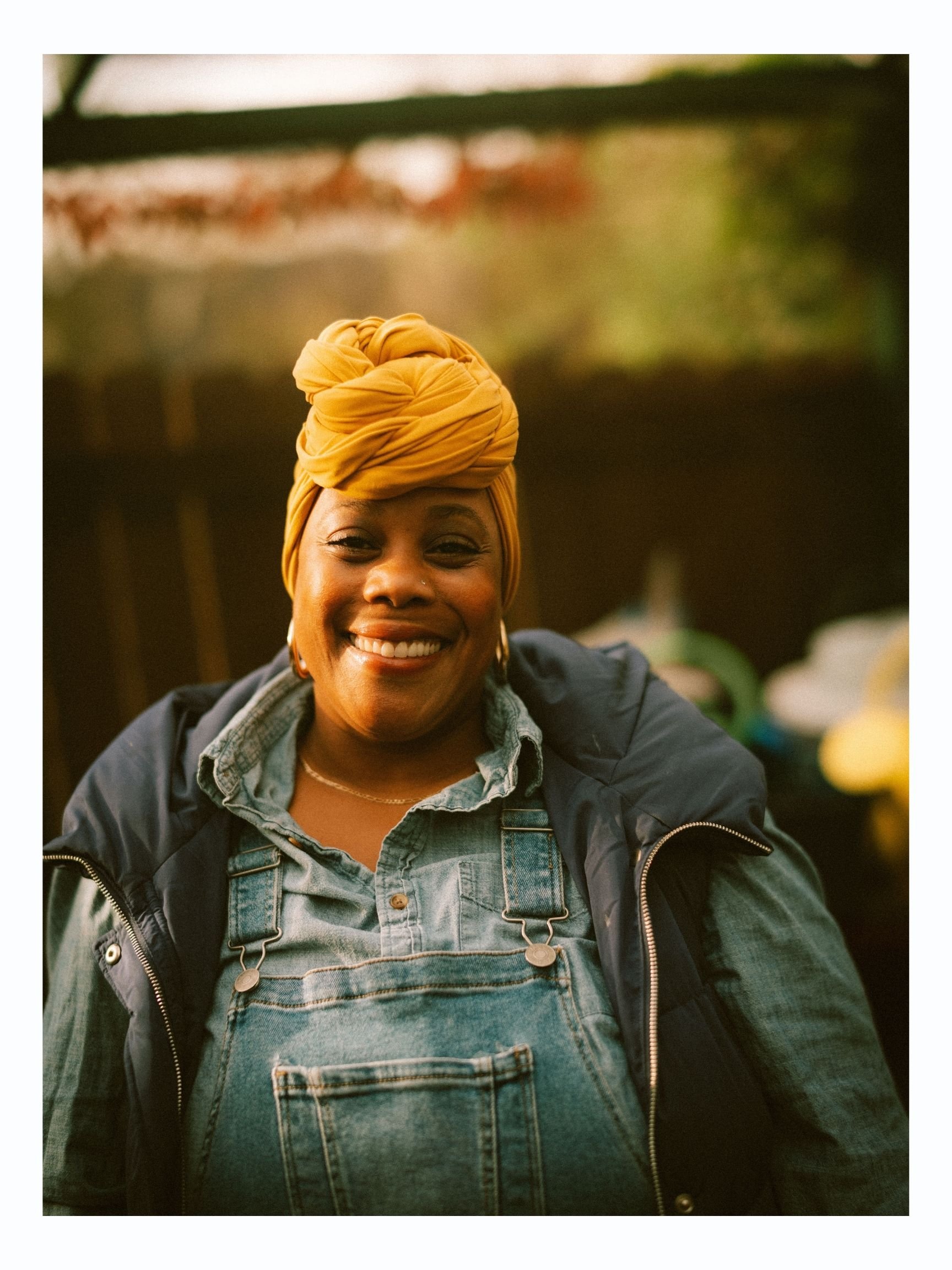 A woman smiling outdoors wearing a colorful headwrap, denim overalls, and a dark jacket.