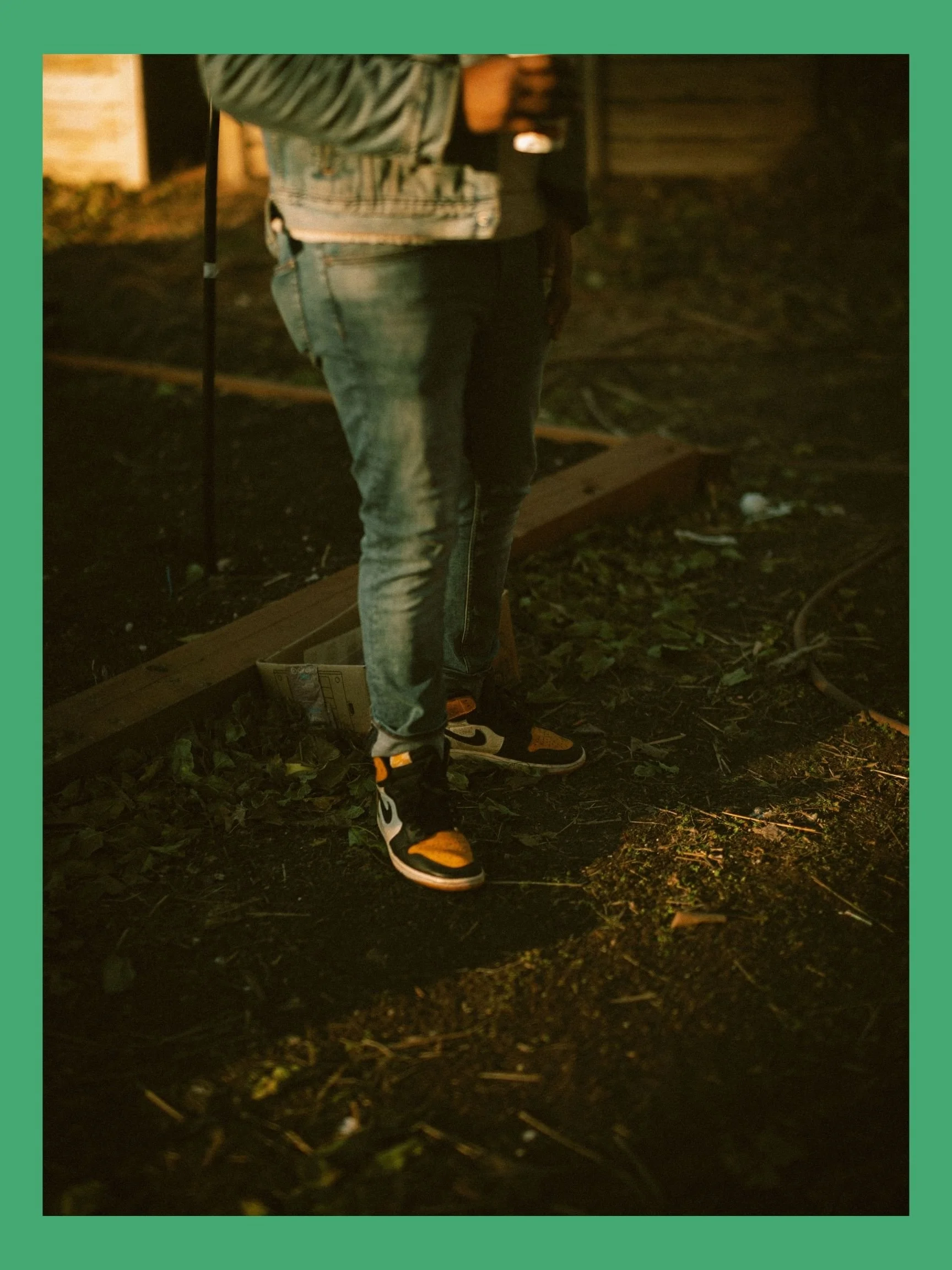 Close-up of a person's lower body wearing jeans and sneakers, standing on outdoor ground with scattered leaves and a cable, with a blurred background at night.
