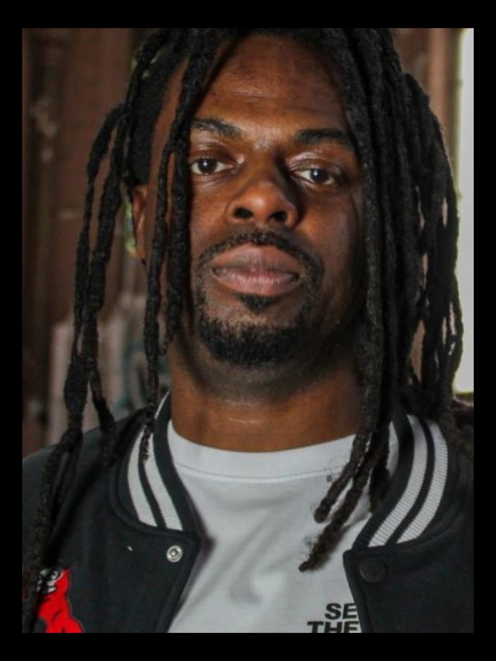 A man with long dreadlocks, facial hair, and wearing a black varsity jacket with a white t-shirt underneath. The background appears to be indoor with wooden elements.