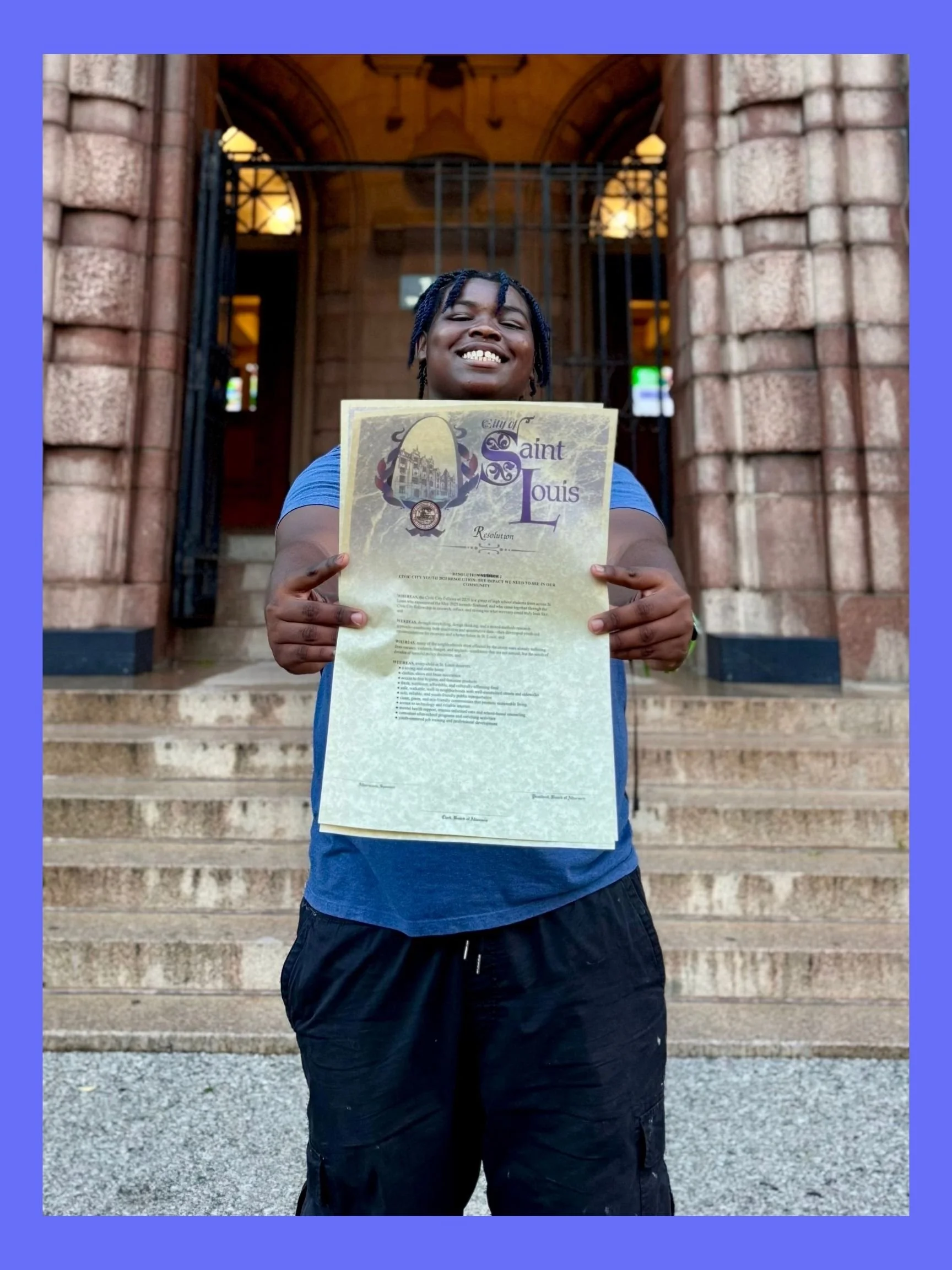A young man with dreadlocks smiling and holding a large document in front of a stone building with steps and arched entrance. The document appears to be a certificate or proclamation from the city of Saint Louis.