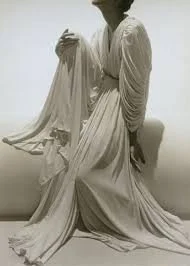 Femme assise, vêtue d’une longue robe blanche drapée et très fluide, dans une pose calme et sculpturale.