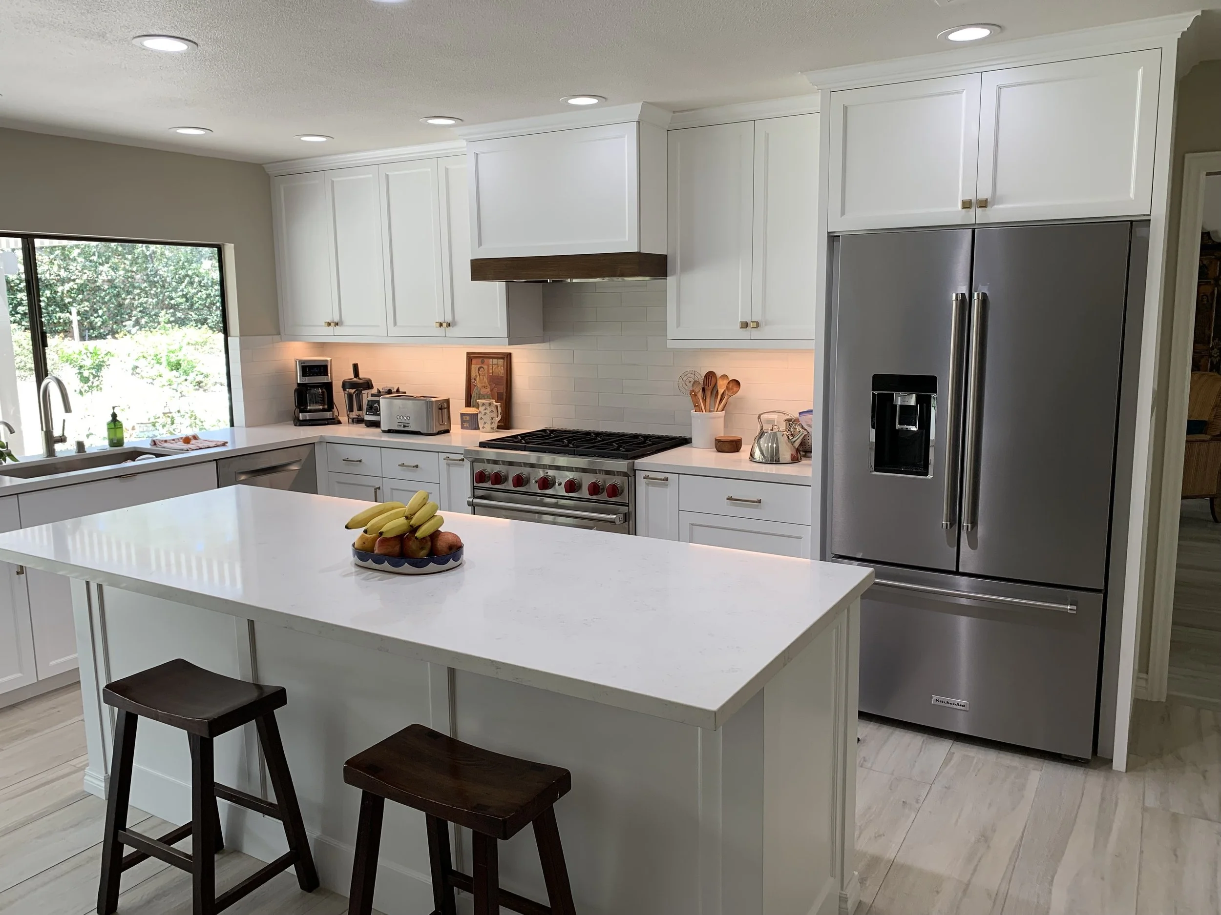 Modern kitchen with white cabinets, a large central island with a fruit bowl, stainless steel refrigerator, and a window overlooking greenery.