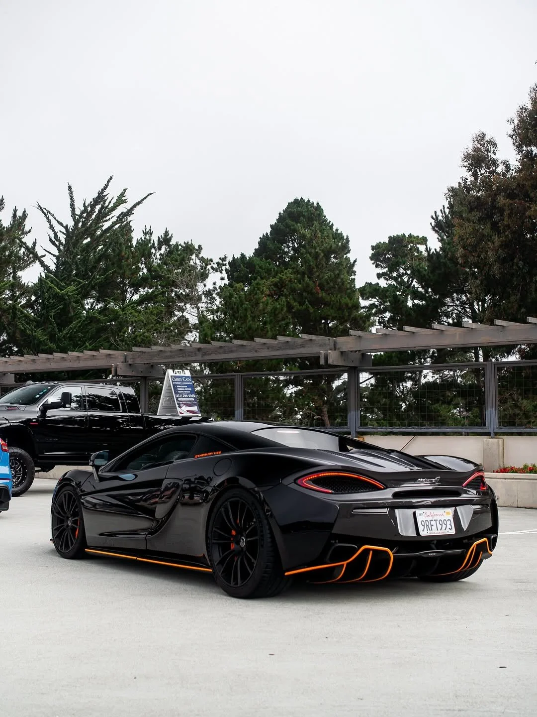 Black sports car with orange accents parked in a parking lot under cloudy sky, with trees in the background.