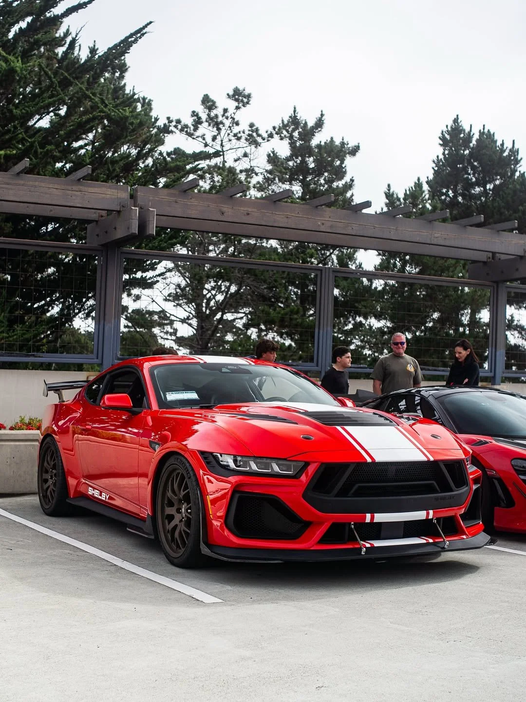 A red sports car with black and white racing stripes parked in a lot, with people standing nearby.
